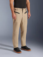Pantalon Alpinestars Flex-AST Canvas, pantalon de moto urbain, marron clair, avec un style workwear héritage, des poches avant zippées et des empiècements ergonomiques aux genoux, porté avec des baskets noires et un t-shirt noir, équipement de moto urbain contemporain