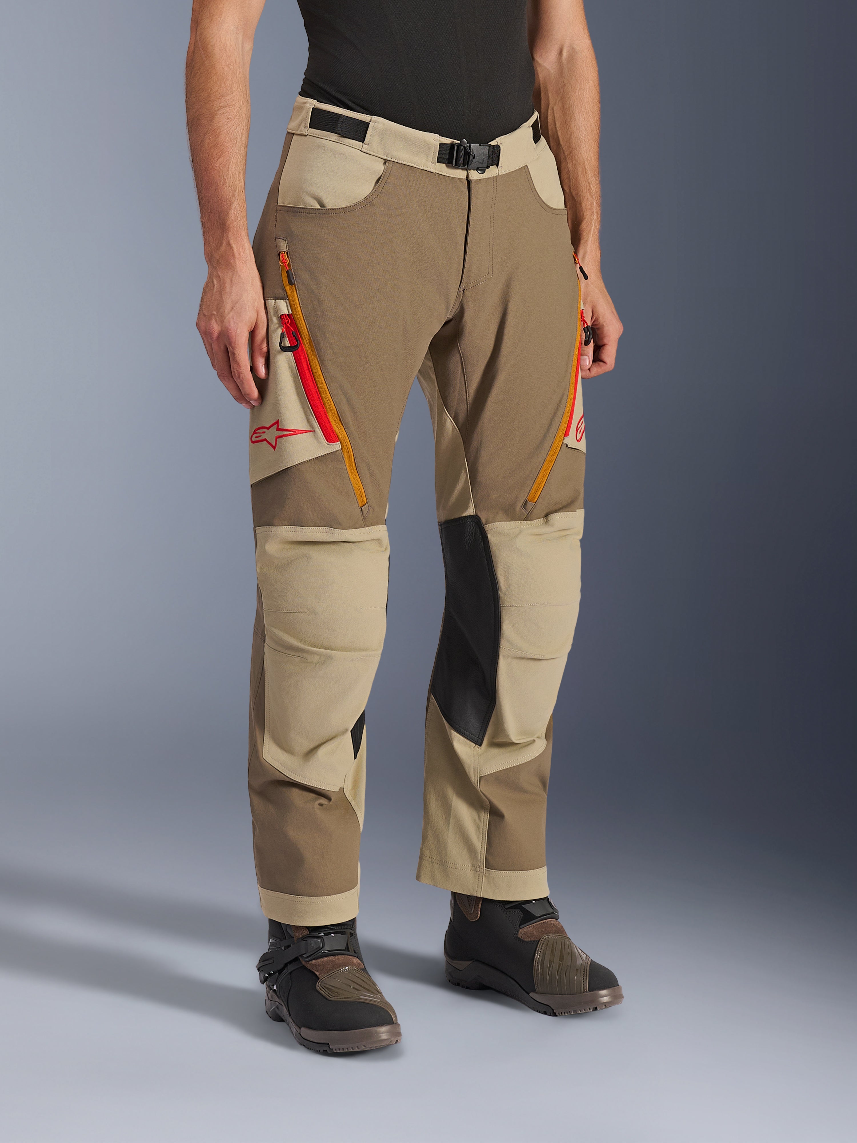 Alpinestars Maxdura Dual Pants, Walnut Aluminum brown and khaki adventure riding pants, featuring orange and red zippered thigh vents, reinforced black leather inner knee panels, and red Astars logo, worn with technical boots.