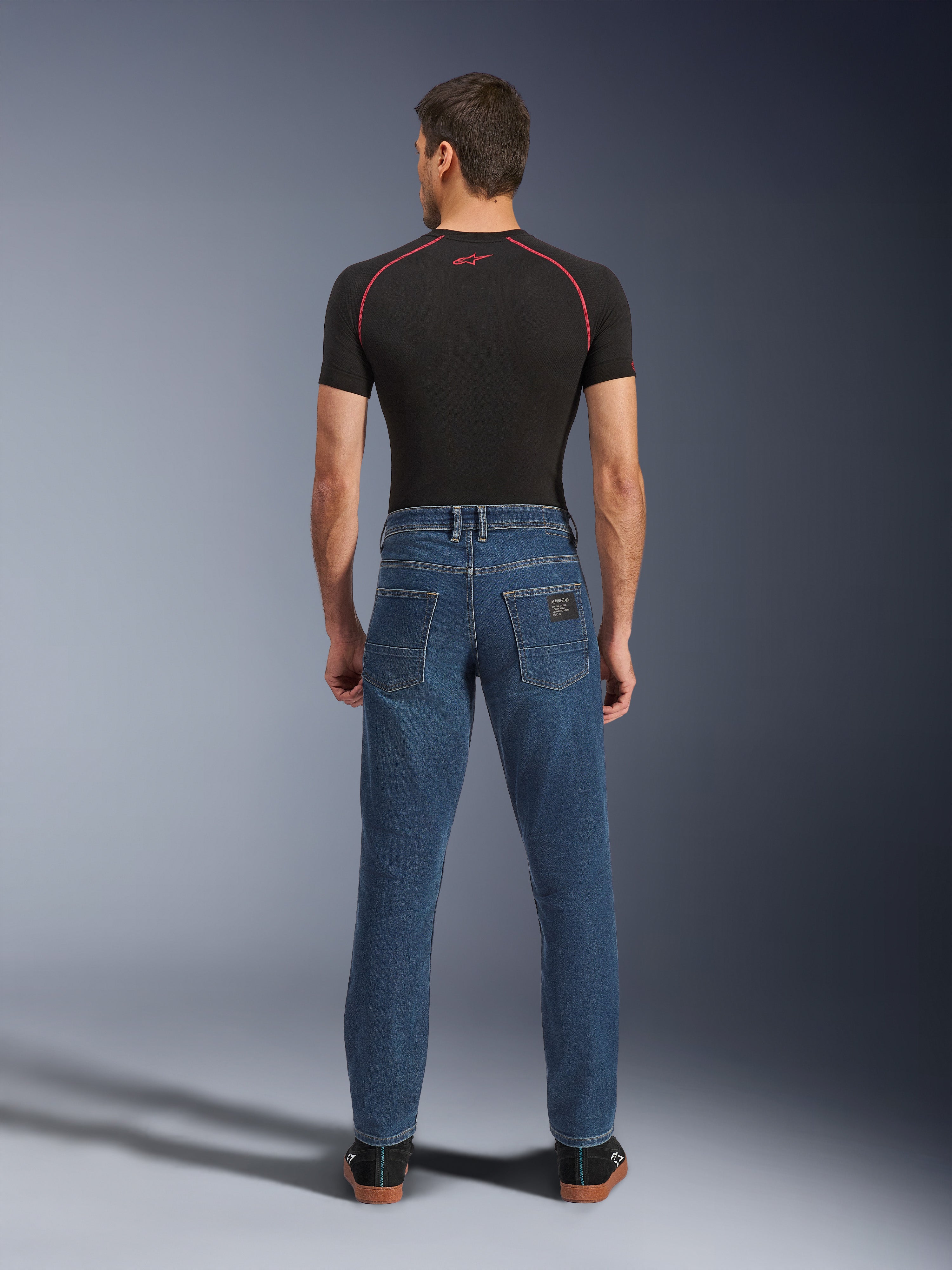 Pantalon en denim Alpinestars Flex-AST 5, bleu Indigo Washed, vue arrière montrant la coupe urbaine fuselée, porté par un mannequin avec un t-shirt technique noir et des baskets, vêtement de protection décontracté pour la moto