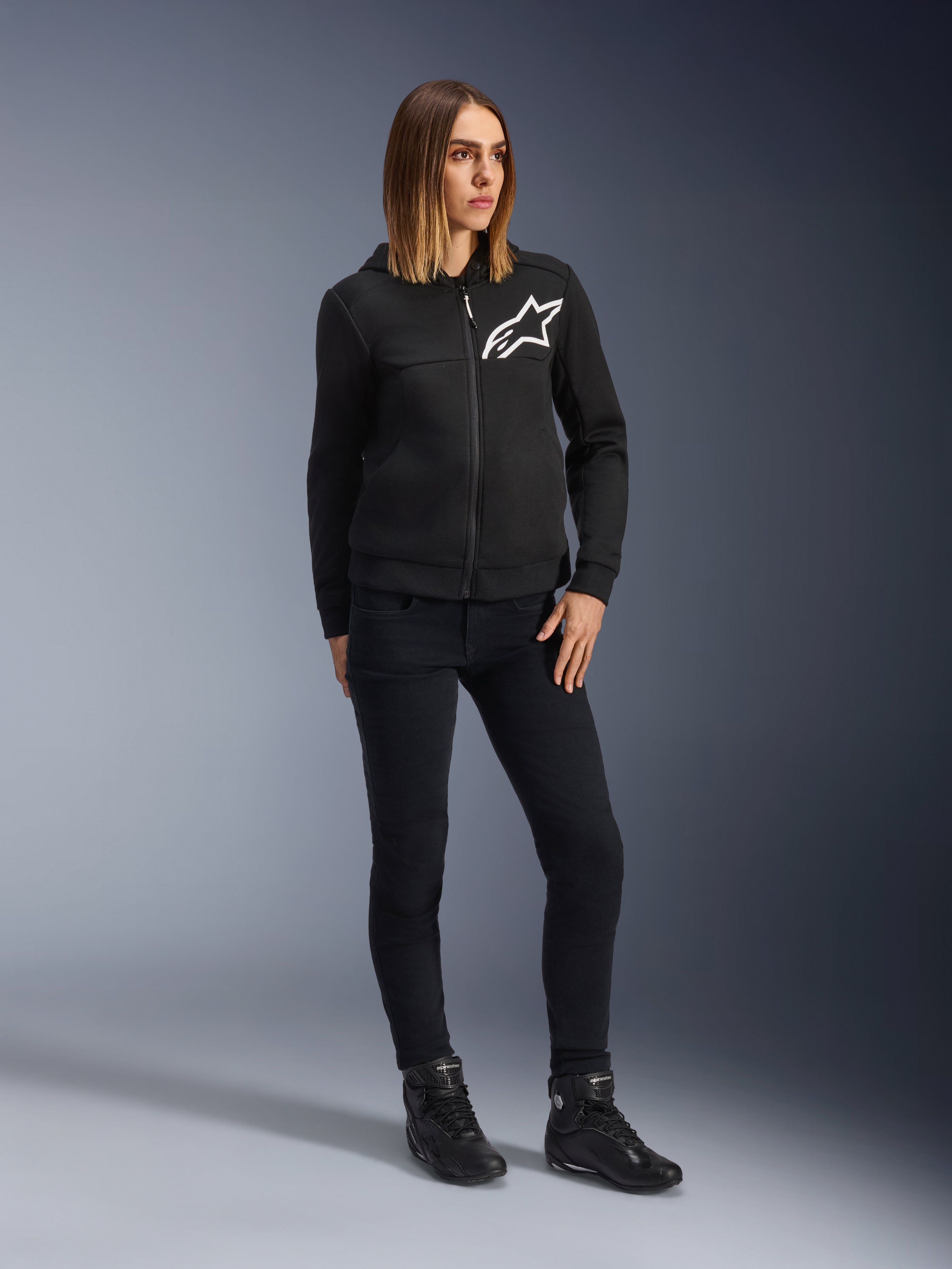 Femme portant le jean de moto Daisy V3 Femme Alpinestars, pantalon de moto coupe slim en Rinse Black, associé à un sweat à capuche zippé noir et des bottes de moto, équipement de protection urbain décontracté