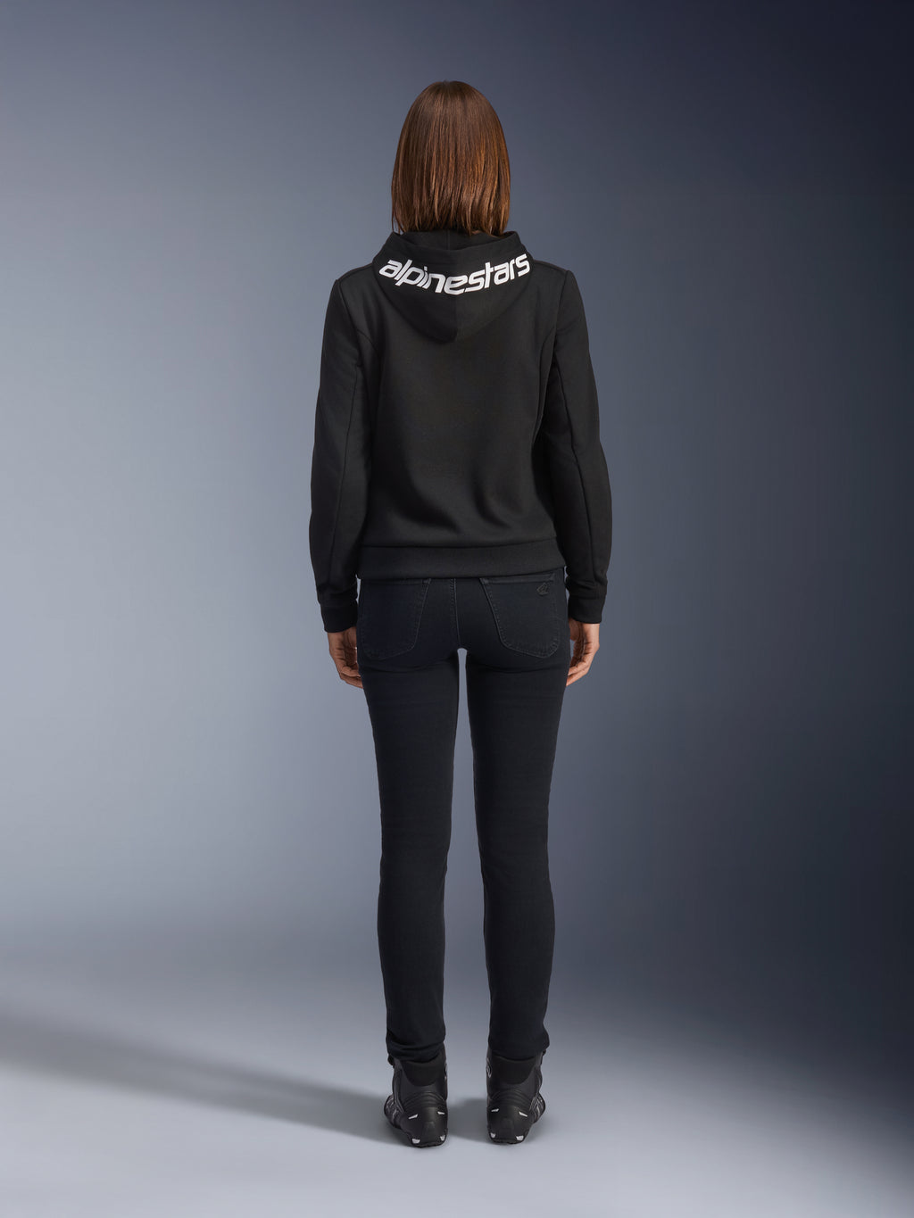 Denim de moto pour femme Daisy V3, Black Rinse, pantalon de moto coupe slim, vue arrière montrant la taille haute et une silhouette féminine, associé à un sweat à capuche Alpinestars noir et des bottes de moto noires, équipement de conduite urbain décontracté