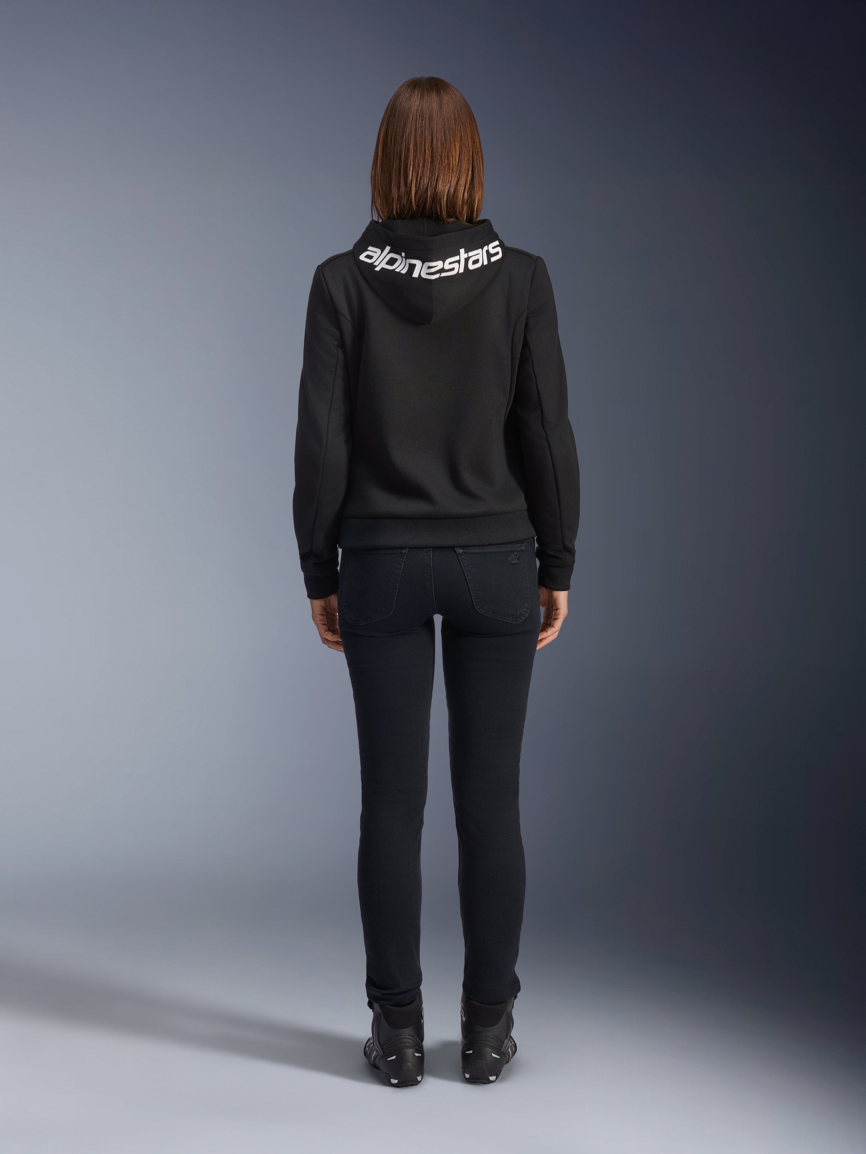 Denim de moto pour femme Daisy V3, Black Rinse, pantalon de moto coupe slim, vue arrière montrant la taille haute et une silhouette féminine, associé à un sweat à capuche Alpinestars noir et des bottes de moto noires, équipement de conduite urbain décontracté