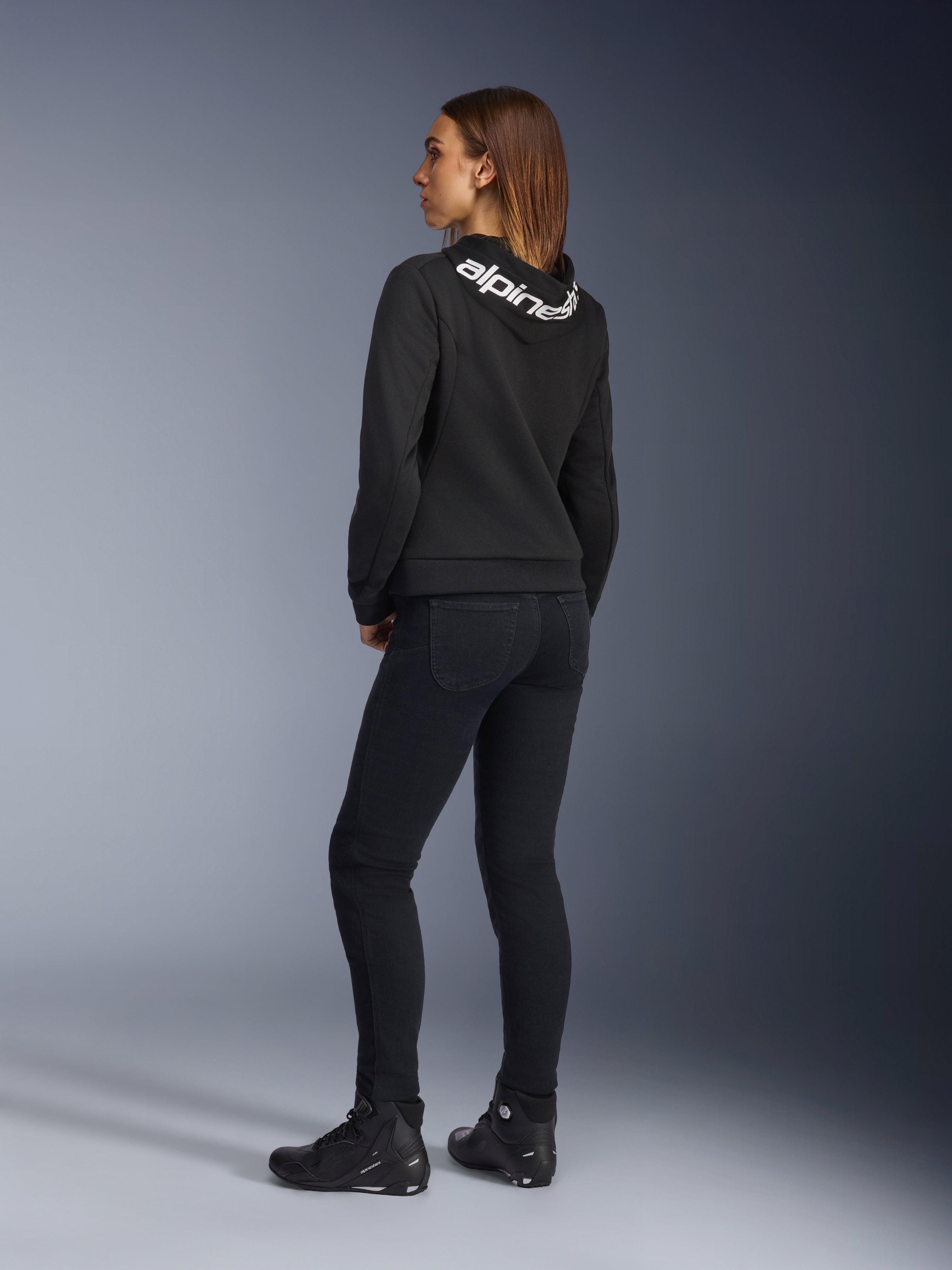 Alpinestars Daisy V3 Women's Riding Denim, pantalon de moto pour femme, noir, vue arrière montrant la coupe ajustée et la taille haute, associé à un sweat à capuche Alpinestars noir et des bottes de pilotage pour un look moto urbain