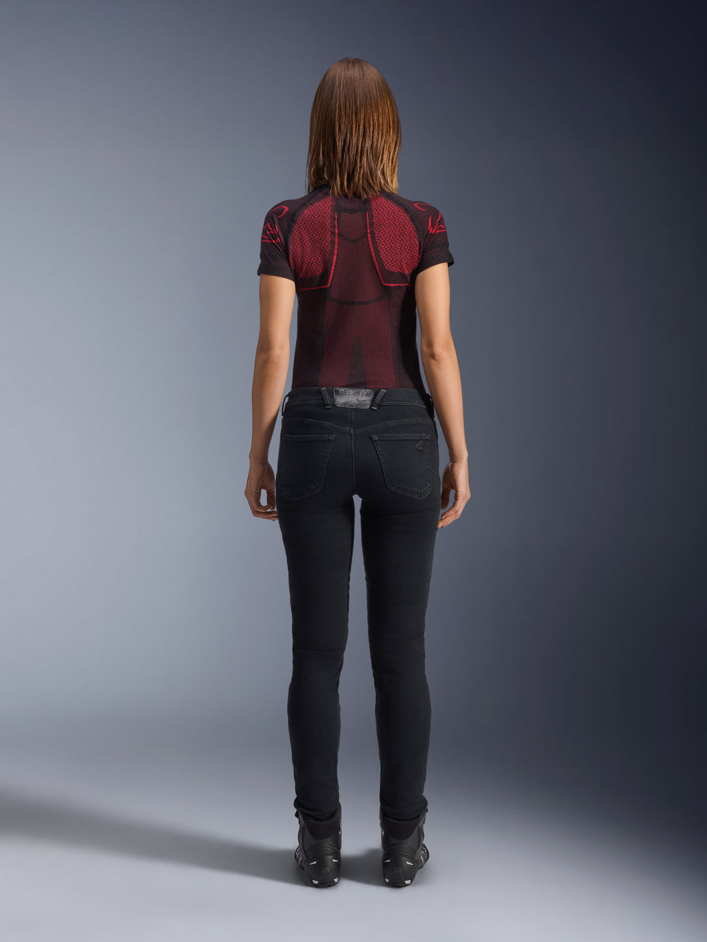 Alpinestars Daisy V3 Women's Riding Denim, Noir Rinse, pantalon de moto coupe slim vu de dos, doté d'une taille haute et de poches arrière, associé à un haut technique à motifs rouge et noir et des bottes noires, équipement de moto urbain pour femme