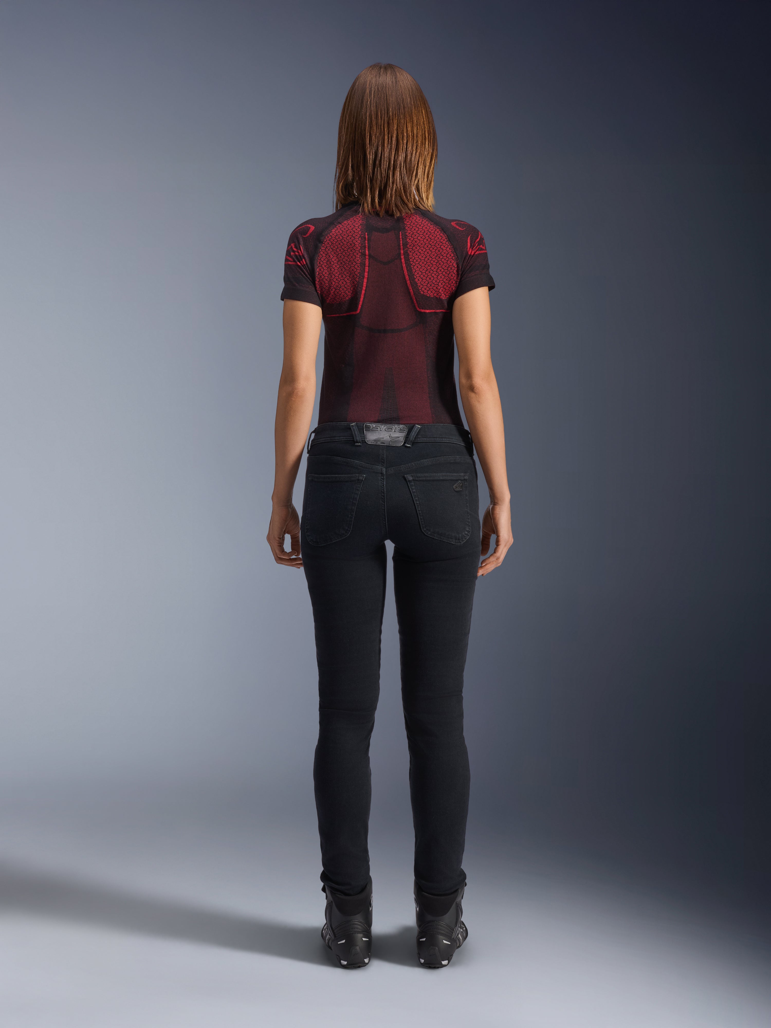 Alpinestars Daisy V3 Women's Riding Denim, Noir Rinse, pantalon de moto coupe slim vu de dos, doté d'une taille haute et de poches arrière, associé à un haut technique à motifs rouge et noir et des bottes noires, équipement de moto urbain pour femme