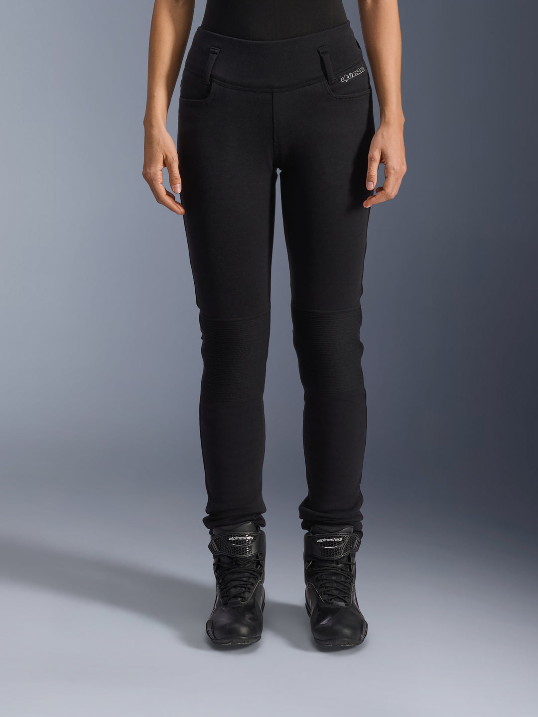 Alpinestars Womens Banshee Leggings, pantalon de moto noir, coupe ajustée avec empiècements en accordéon aux genoux, porté avec des bottes de moto montantes noires, équipement de route urbain au style décontracté