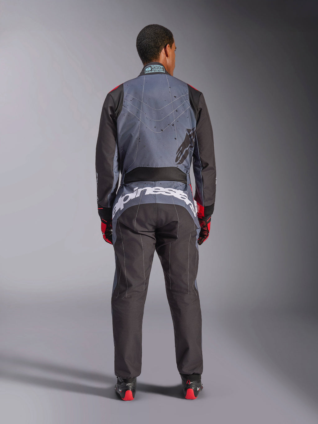 Alpinestars KMX-9 V3 Graphic 6 Suit, Karting Suit, Anthracite Black and Red, rear view showing large white Alpinestars branding across the lower back, gray upper body with black splatter graphics, black arms and legs, worn with red and black racing gloves and boots.