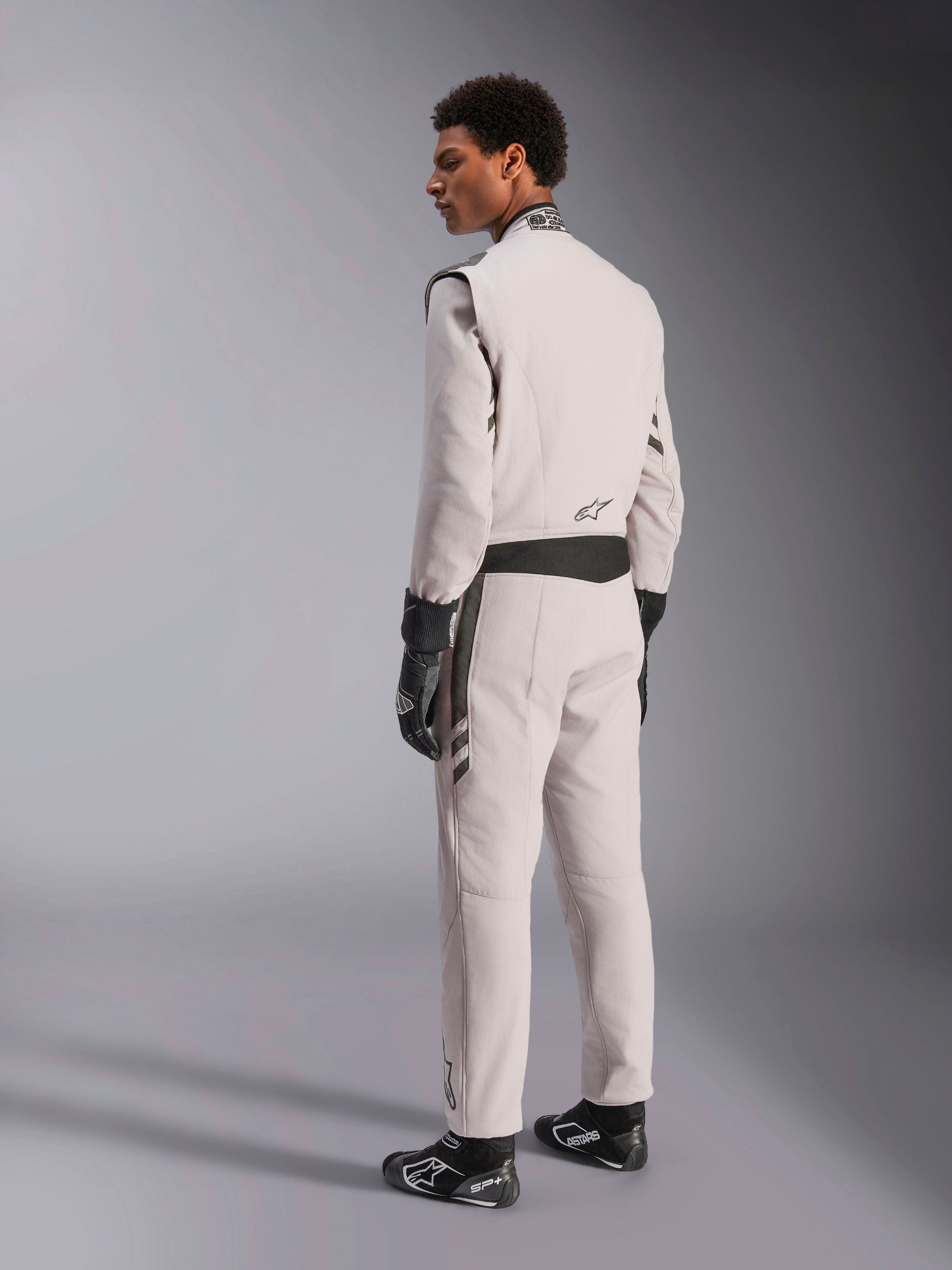 Alpinestars GP Race V3 Suit FIA, combinaison de course Argent Anthracite, vue de profil et de dos, portée avec des gants et des bottines de course noirs, avec des détails gris foncé à la taille et aux épaules, équipement de protection professionnel pour la course automobile.