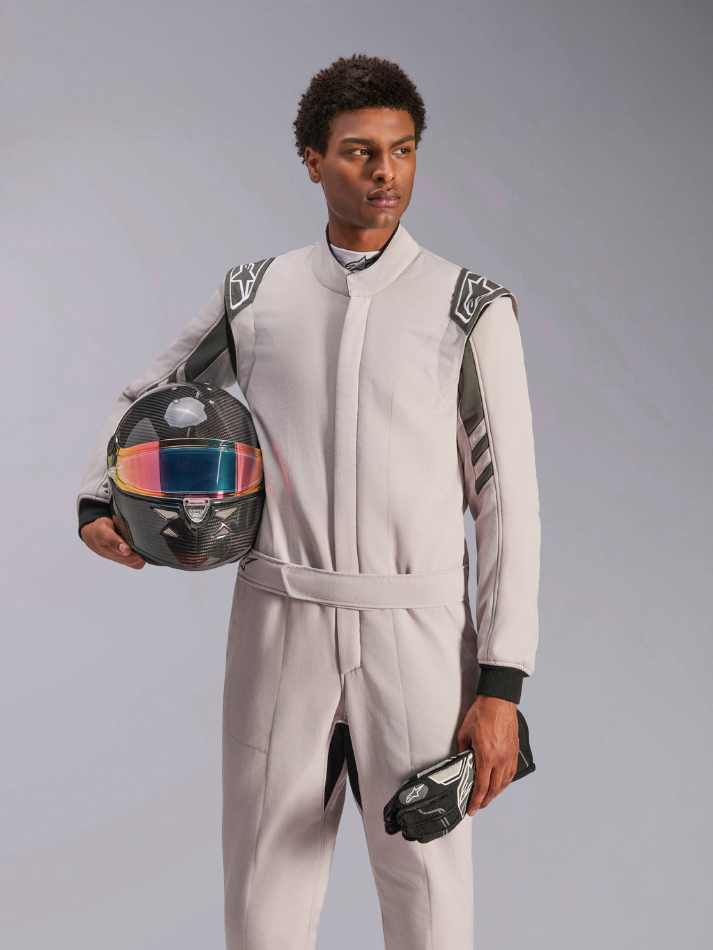 Alpinestars GP Race V3 Suit FIA, combinaison de course automobile, Silver Anthracite, portée par un mannequin tenant un casque en fibre de carbone avec visière irisée et gants de course noirs, équipement de protection de sport automobile professionnel avec épaulettes visibles.