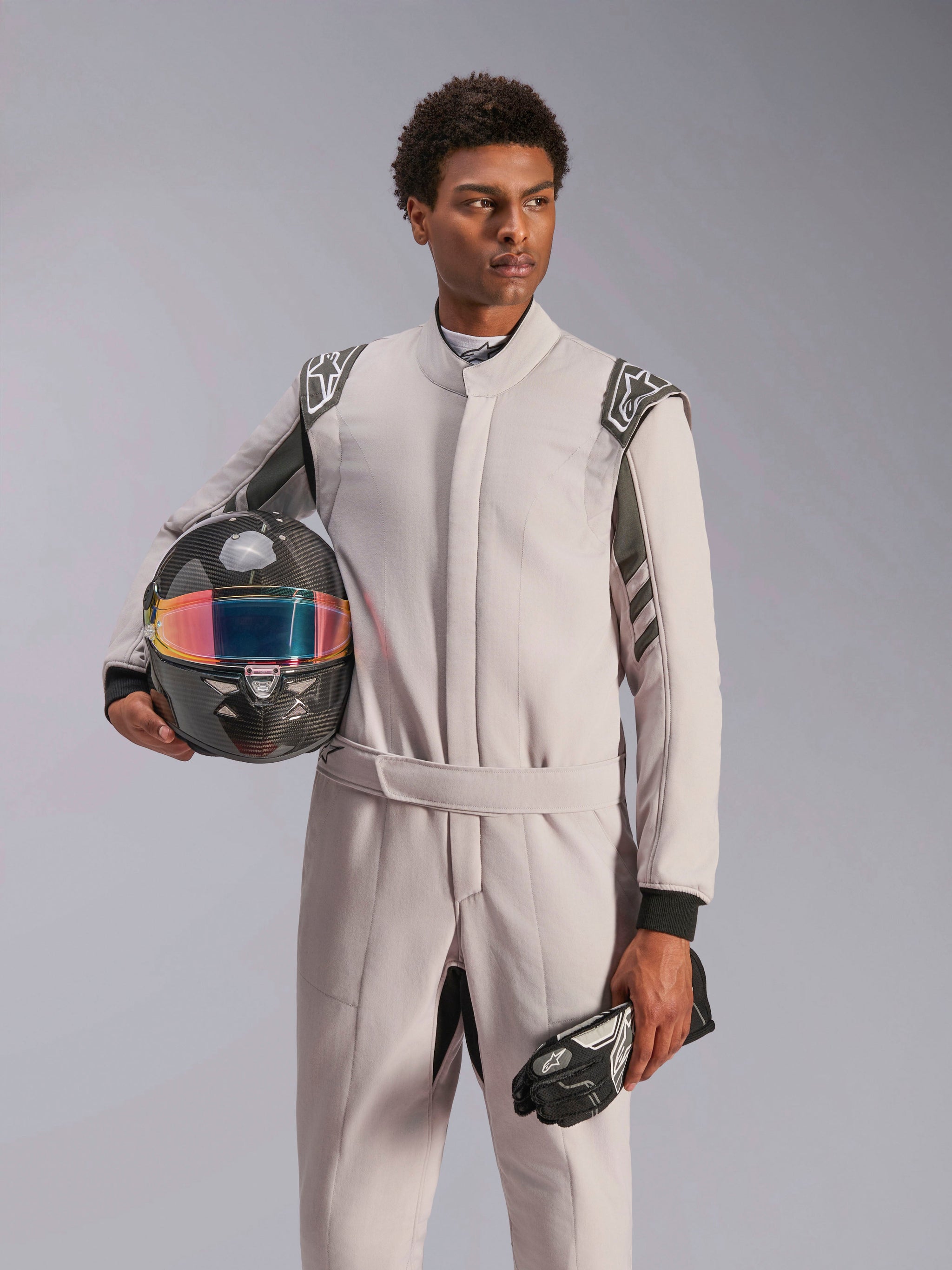 Alpinestars GP Race V3 Suit FIA, combinaison de course automobile, Silver Anthracite, portée par un mannequin tenant un casque en fibre de carbone avec visière irisée et gants de course noirs, équipement de protection de sport automobile professionnel avec épaulettes visibles.