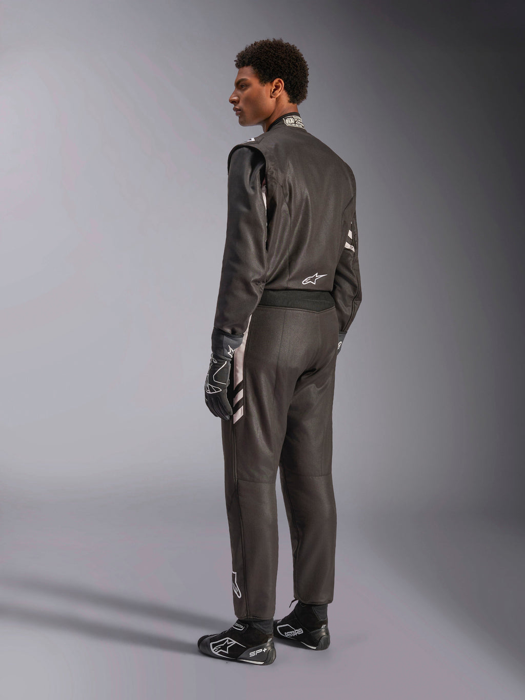 Alpinestars GP Race V3 Suit FIA, combinaison de course automobile noire avec des accents argentés, portée par un mannequin en vue de trois-quarts arrière, associée à des gants et des bottes de course noirs assortis, équipement de sport automobile professionnel présentant une conception élégante à deux couches.