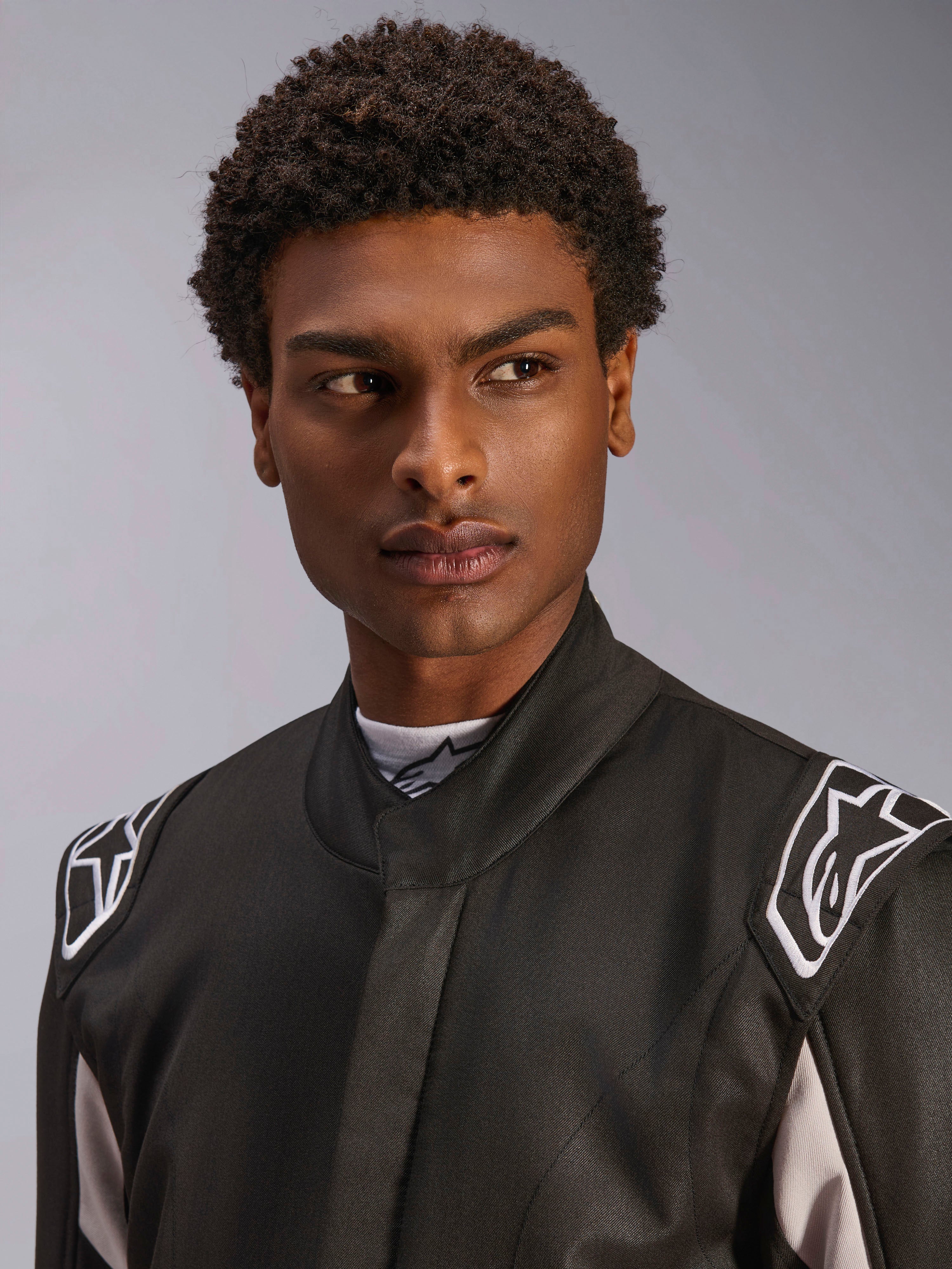 Combinaison Alpinestars GP Race V3 FIA, combinaison de course automobile noire portée par un homme, vue rapprochée montrant le col souple et les logos d'étoiles blancs sur les épaulettes, équipement de protection professionnel pour le sport automobile