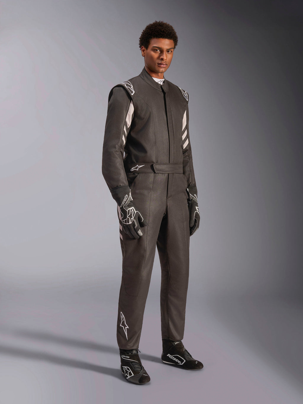 Pilote portant une combinaison Alpinestars GP Race V3 FIA, combinaison de course automobile noir et argent, avec gants et chaussures assortis, détails argentés sur les épaules et le bas des jambes, équipement de course automobile professionnel avec coupe ajustée