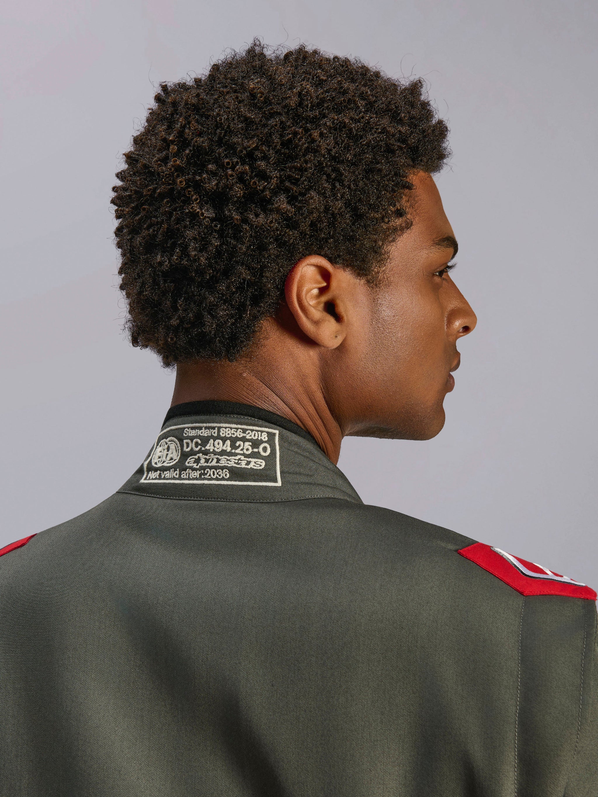 Alpinestars GP Race V3 Suit FIA, combinaison de course rouge anthracite, gros plan sur le col arrière montrant l'étiquette de certification FIA 8856-2018 et les épaulettes rouges, vue détaillée d'un équipement de sport automobile professionnel.