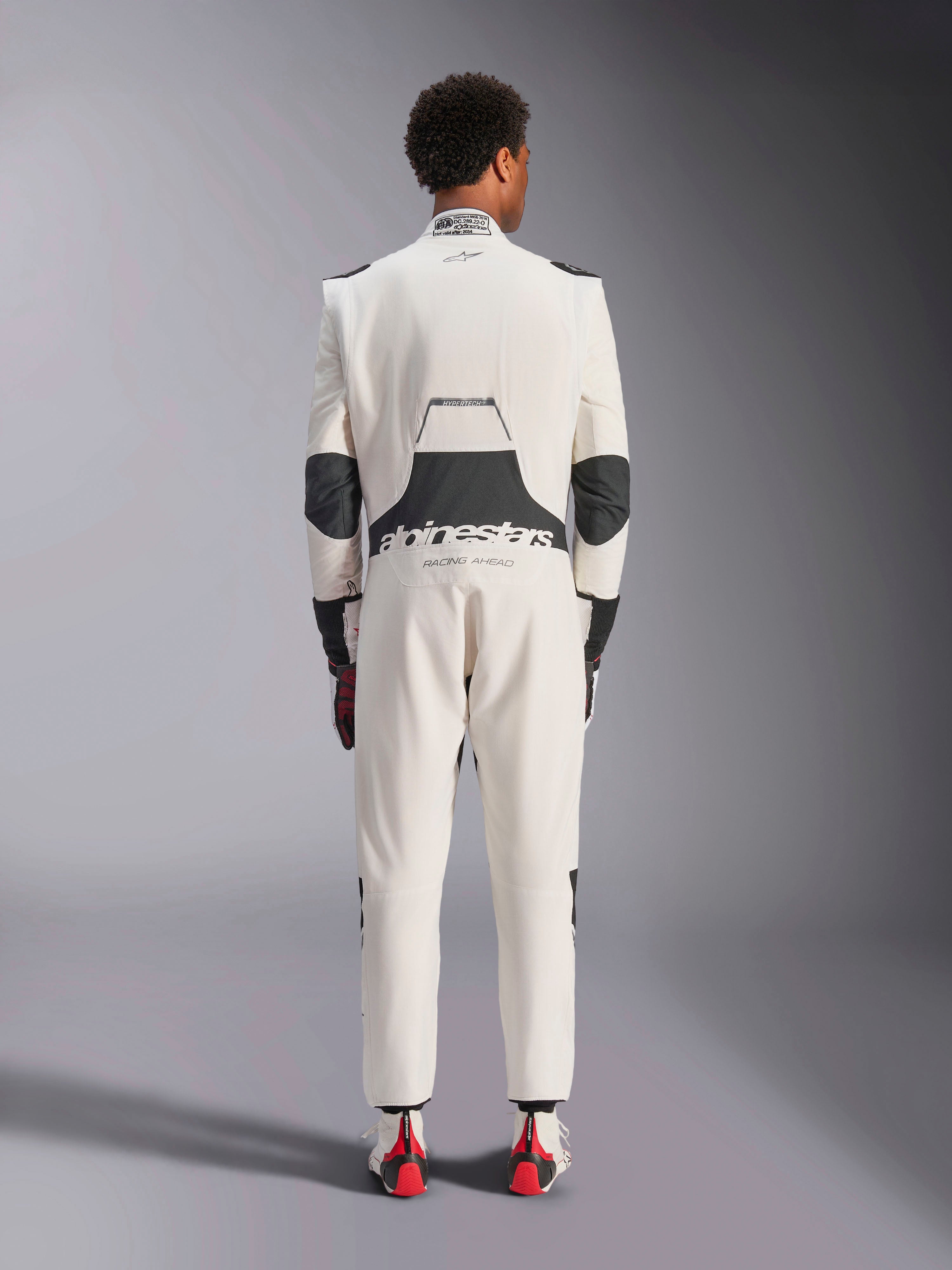 Vue arrière d'un mannequin portant la combinaison Alpinestars Hypertech V3 en Blanc Noir Argent, montrant le large panneau stretch lombaire noir avec les logos « alpinestars » et « RACING AHEAD », des inserts noirs aux coudes, l'étiquette d'homologation FIA sur le col, et une coupe athlétique slim conçue pour la course professionnelle.