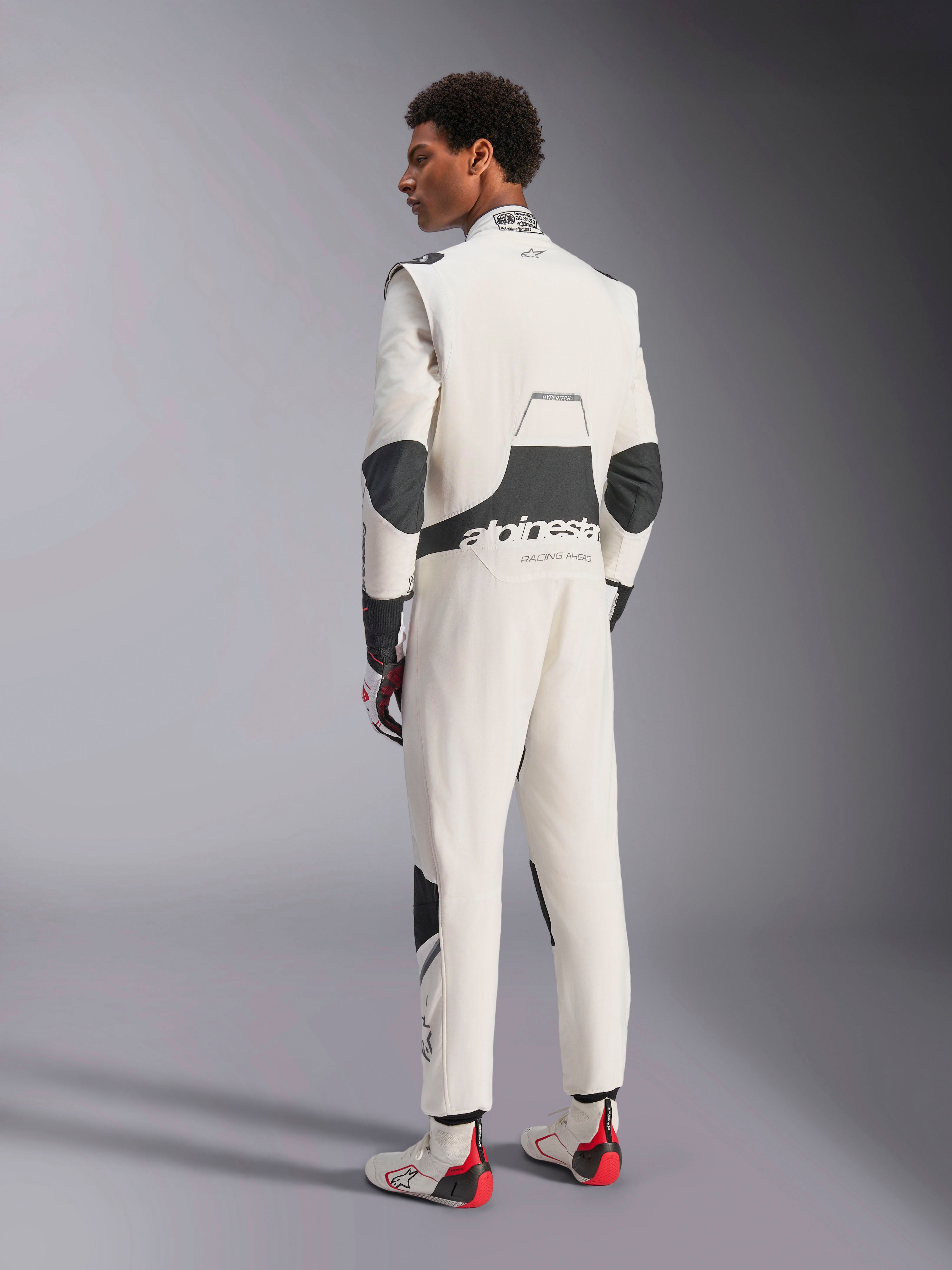 Combinaison Alpinestars Hypertech V3, combinaison de course automobile professionnelle, blanc, noir et argent, vue arrière du modèle montrant des panneaux extensibles noirs sur les lombaires et les coudes, logo Alpinestars dans le dos, avec gants et bottes de course assortis pour un look complet de sport automobile