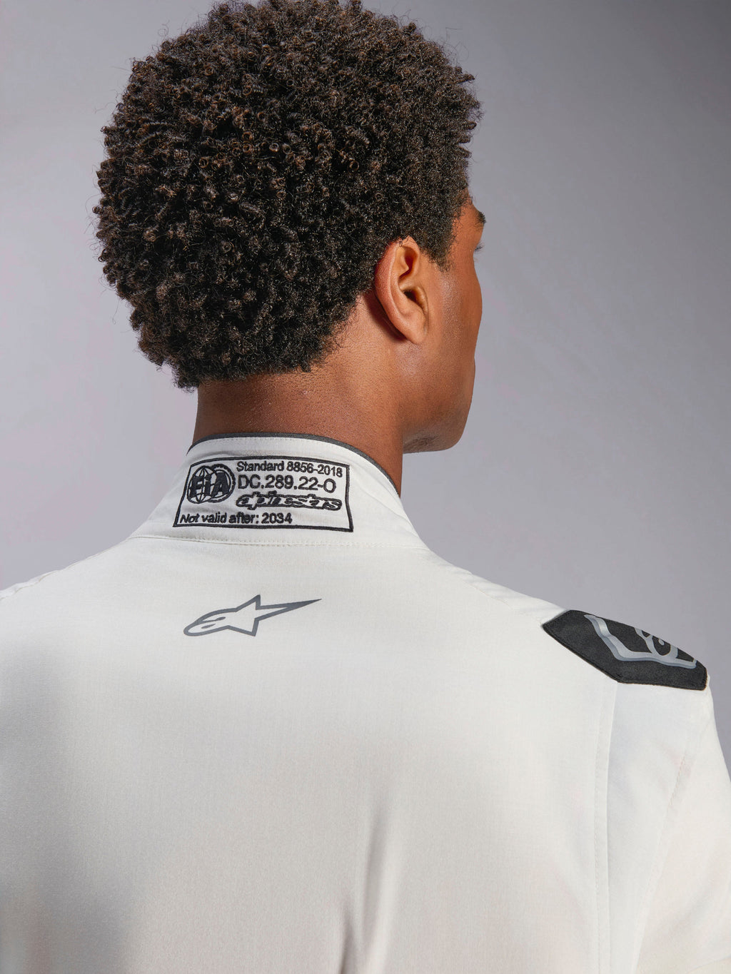 Combinaison Alpinestars Hypertech V3, blanc, vue détaillée du col arrière et de l'épaule, présentant l'étiquette de certification FIA 8856-2018, le logo Alpinestars gris imprimé et la poignée d'extraction noire, équipement de course professionnel
