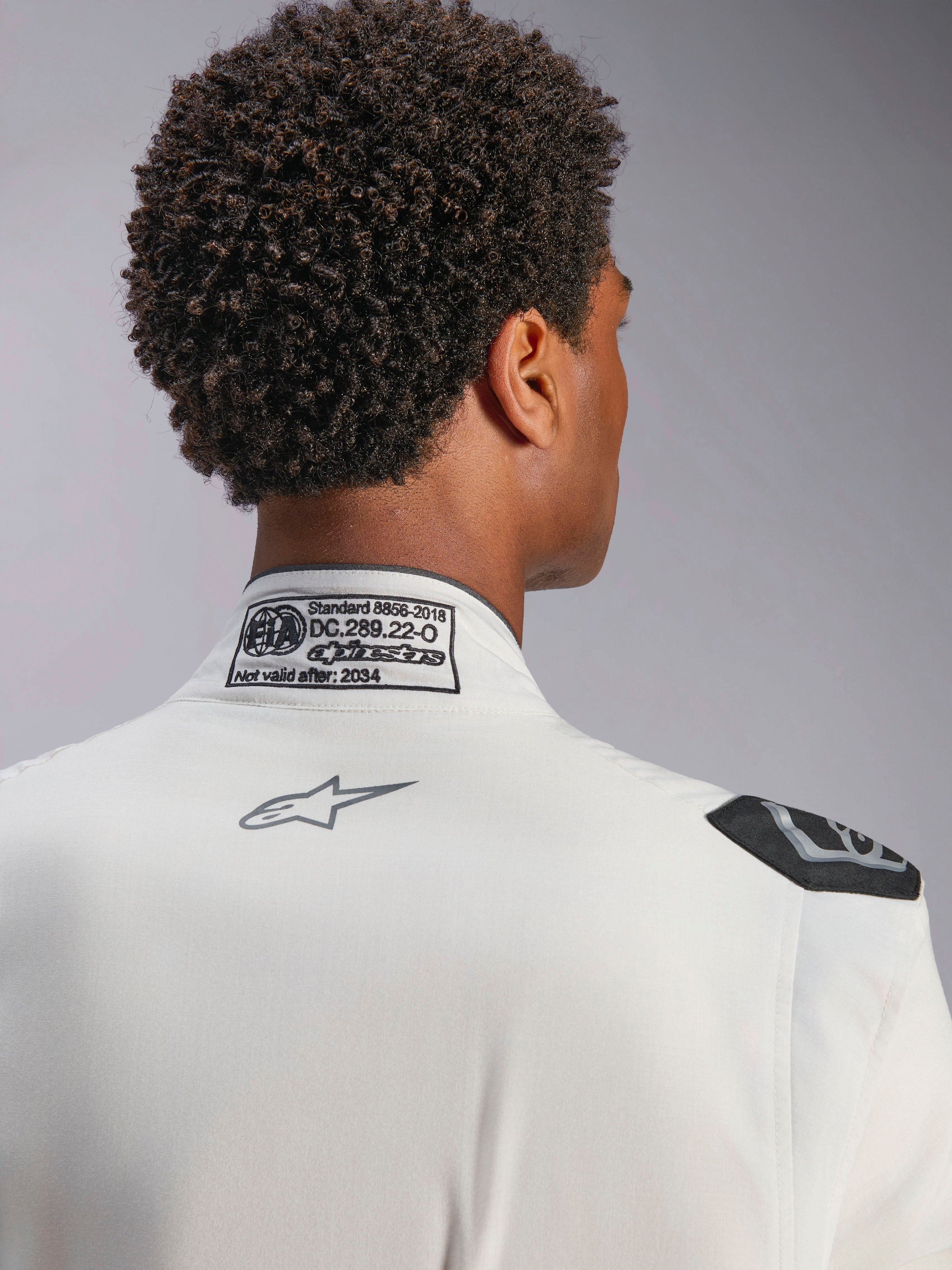 Combinaison Alpinestars Hypertech V3, blanc, vue détaillée du col arrière et de l'épaule, présentant l'étiquette de certification FIA 8856-2018, le logo Alpinestars gris imprimé et la poignée d'extraction noire, équipement de course professionnel