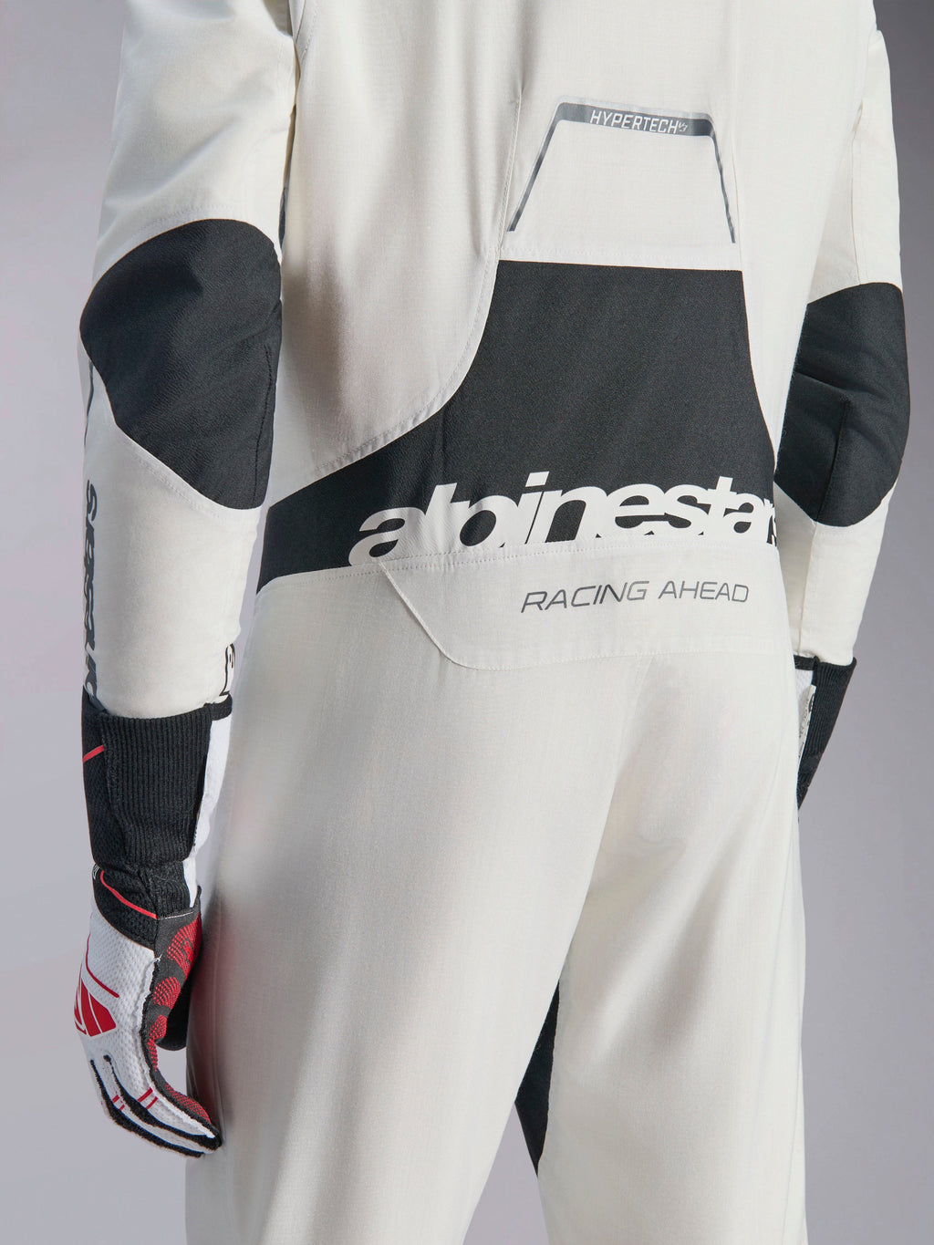 Combinaison Alpinestars Hypertech V3, combinaison de course automobile blanc, noir et argent, vue arrière montrant le grand logo 'alpinestars' noir et le texte 'RACING AHEAD', avec des panneaux extensibles noirs aux coudes et aux lombaires, vêtement de protection professionnel pour le sport automobile.