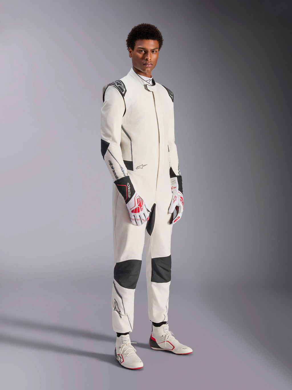 Mannequin homme portant la combinaison Alpinestars Hypertech V3, blanche avec des accents noirs et argentés, équipement de course automobile professionnel doté de panneaux noirs sur les épaules et les genoux, assorti de gants de course blancs et rouges et de bottines de course blanches