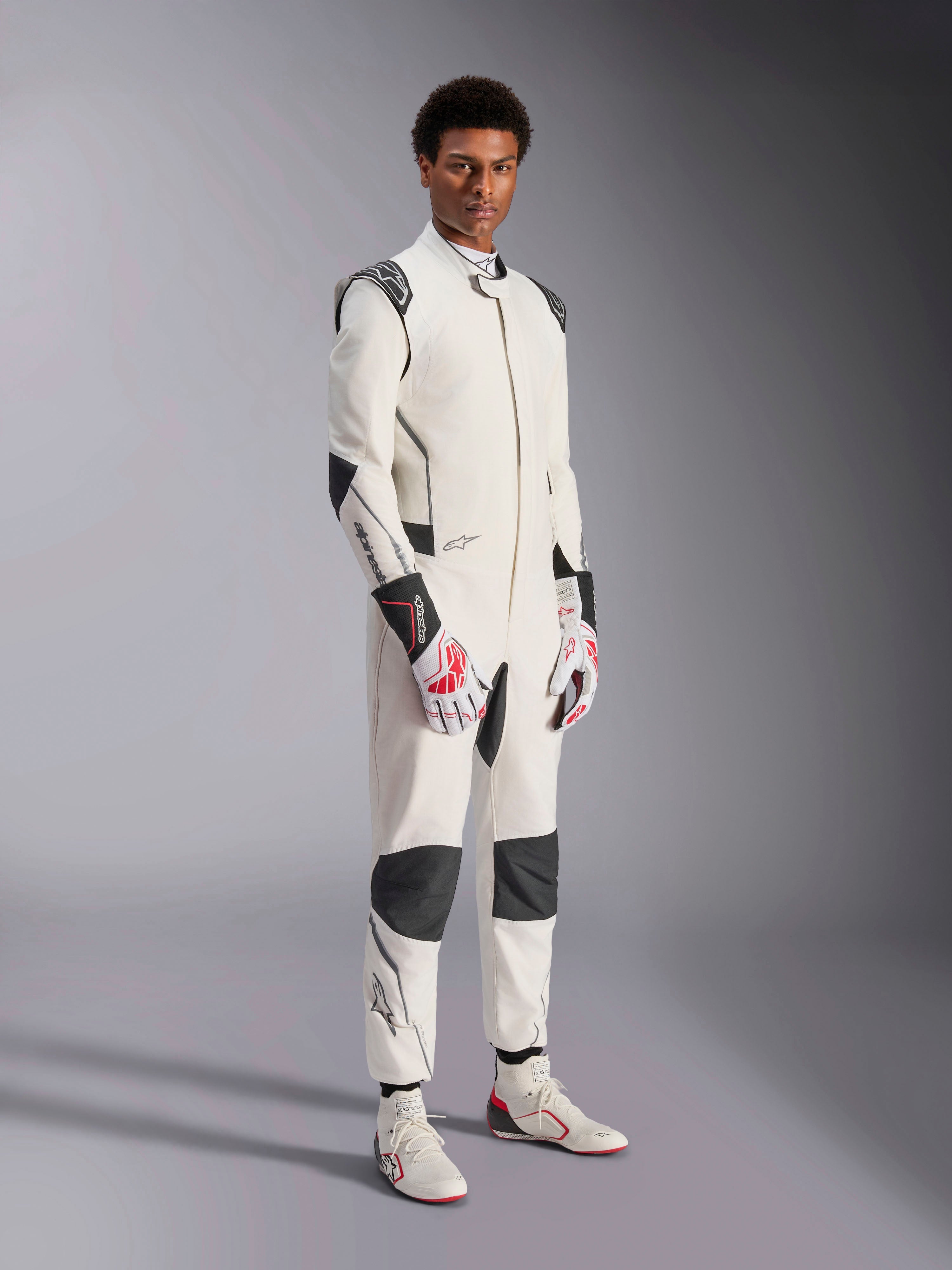 Mannequin homme portant la combinaison Alpinestars Hypertech V3, blanche avec des accents noirs et argentés, équipement de course automobile professionnel doté de panneaux noirs sur les épaules et les genoux, assorti de gants de course blancs et rouges et de bottines de course blanches