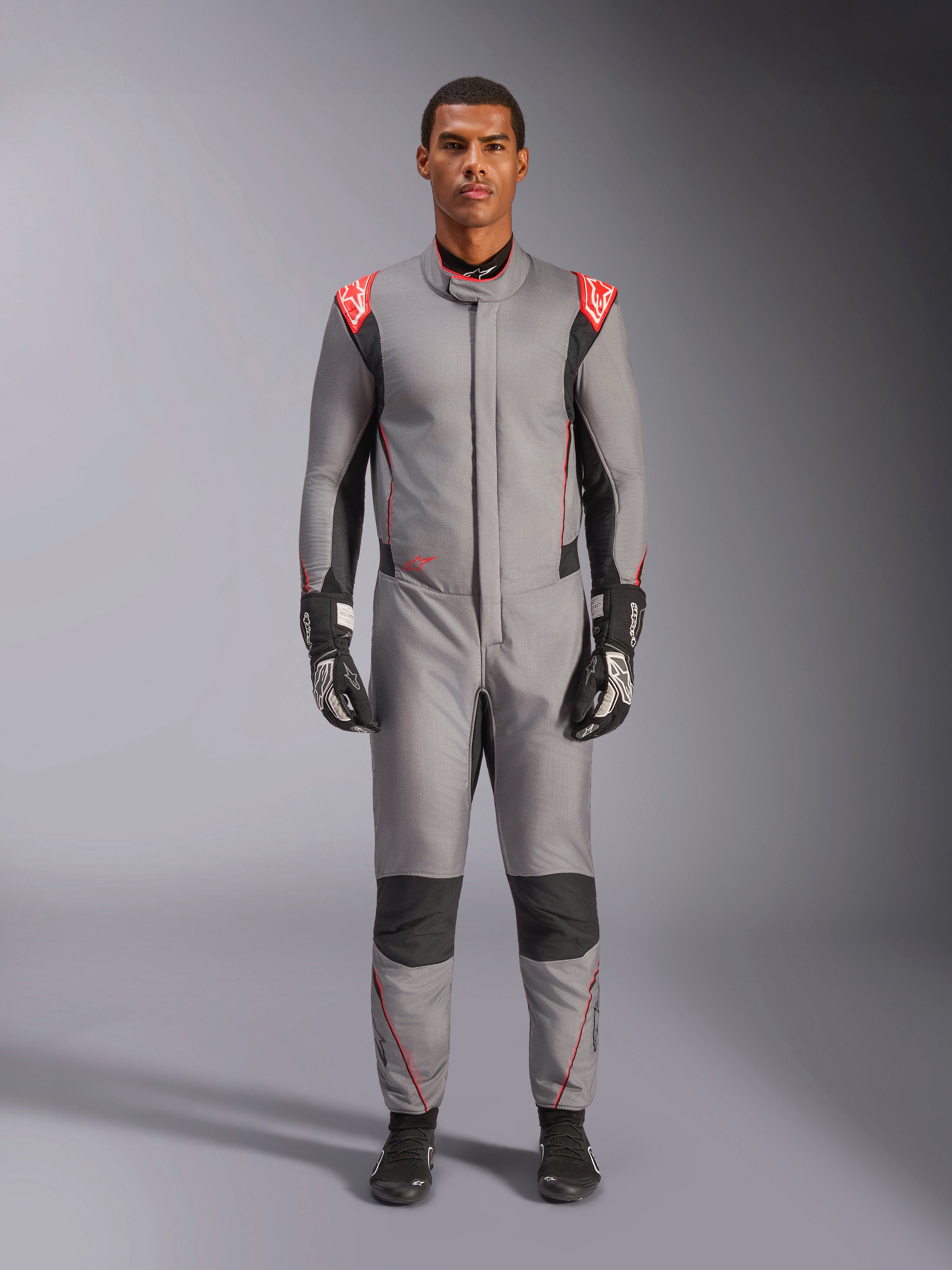 Alpinestars Hypertech V3 Suit, combinaison de course automobile professionnelle, gris moyen, noir et rouge fluo, portée par un mannequin avec gants et chaussures de course noirs, coupe athlétique slim, détails rouge fluo aux épaules et empiècements noirs aux genoux et sur les côtés.