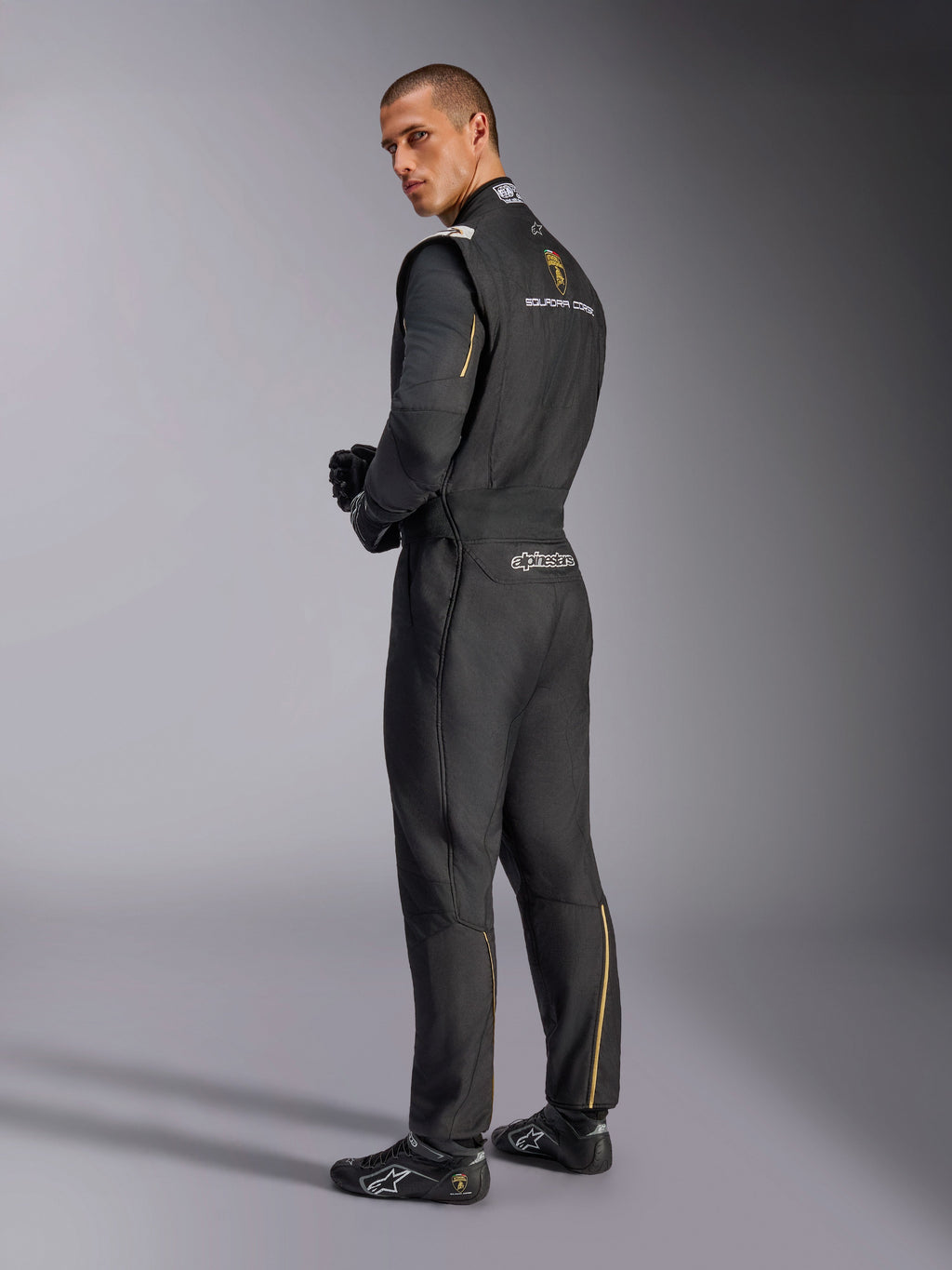 Combinaison Alpinestars GP Tech V4 Édition Limitée Lamborghini, combinaison de course automobile professionnelle, noire avec accents argentés et dorés, vue de dos montrant les logos Lamborghini Squadra Corse et Alpinestars, portée avec des gants et des chaussures de course noirs, design sport automobile épuré
