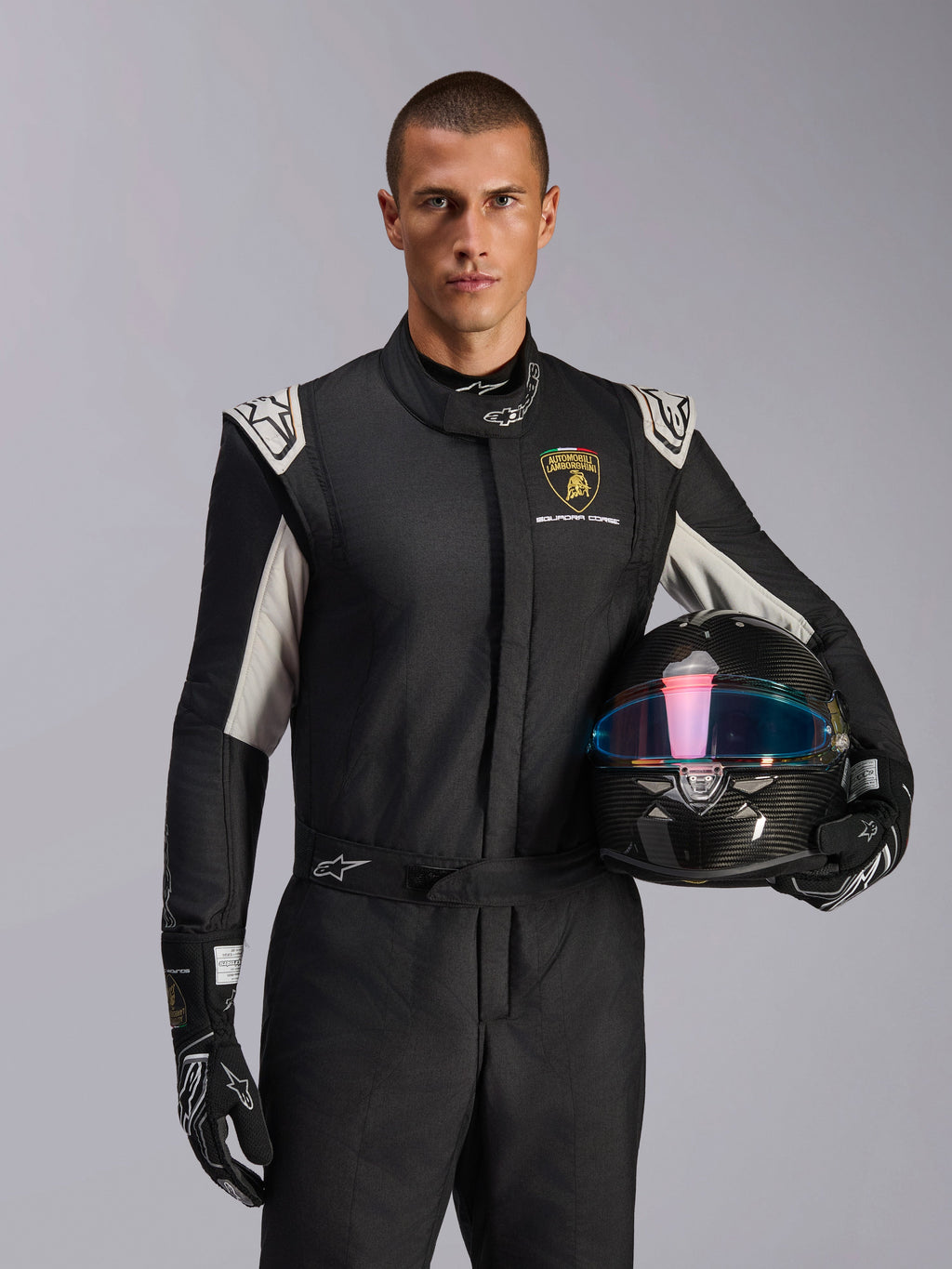 Modèle masculin portant la combinaison Limited Edition Lamborghini GP Tech V4, combinaison de course noire avec accents argentés et logo Lamborghini Squadra Corse, tenant un casque en fibre de carbone, gants de course noirs avec logos blancs, équipement de course automobile professionnel