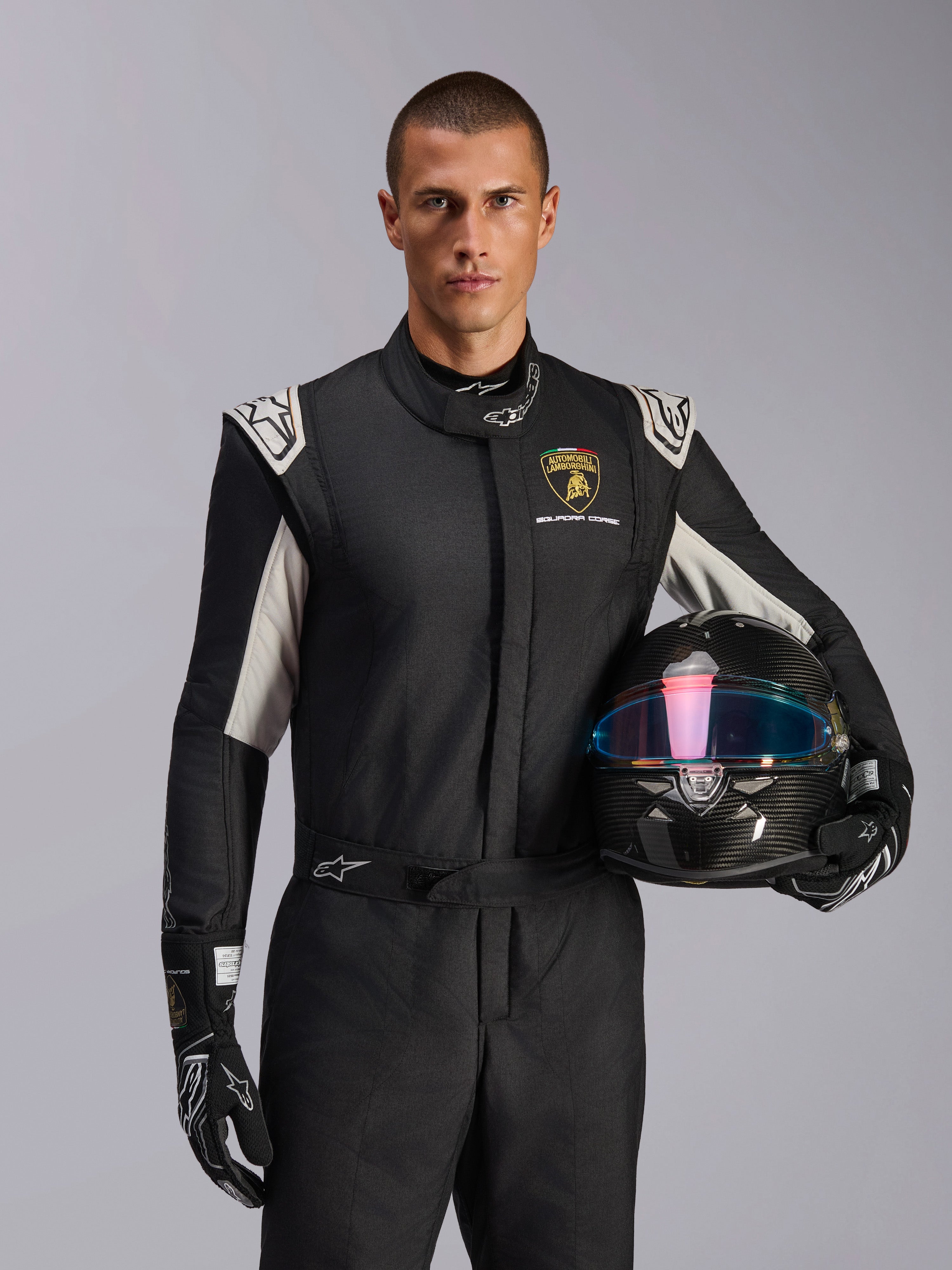 Combinaison Alpinestars GP Tech V4 Édition Limitée Lamborghini, combinaison de course automobile, noir, argent et or, portée par un mannequin tenant un casque en fibre de carbone avec une visière irisée, avec logo Lamborghini Squadra Corse et accents blancs sur les épaules