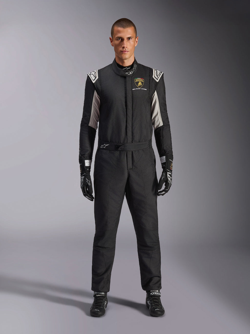 Alpinestars Édition Limitée Lamborghini GP Tech V4 Combinaison FIA, combinaison de course automobile professionnelle, noir avec accents argentés et dorés, arborant le logo Automobili Lamborghini Squadra Corse sur la poitrine, portée avec gants et bottes de course assortis, design athlétique épuré pour la compétition sur circuit