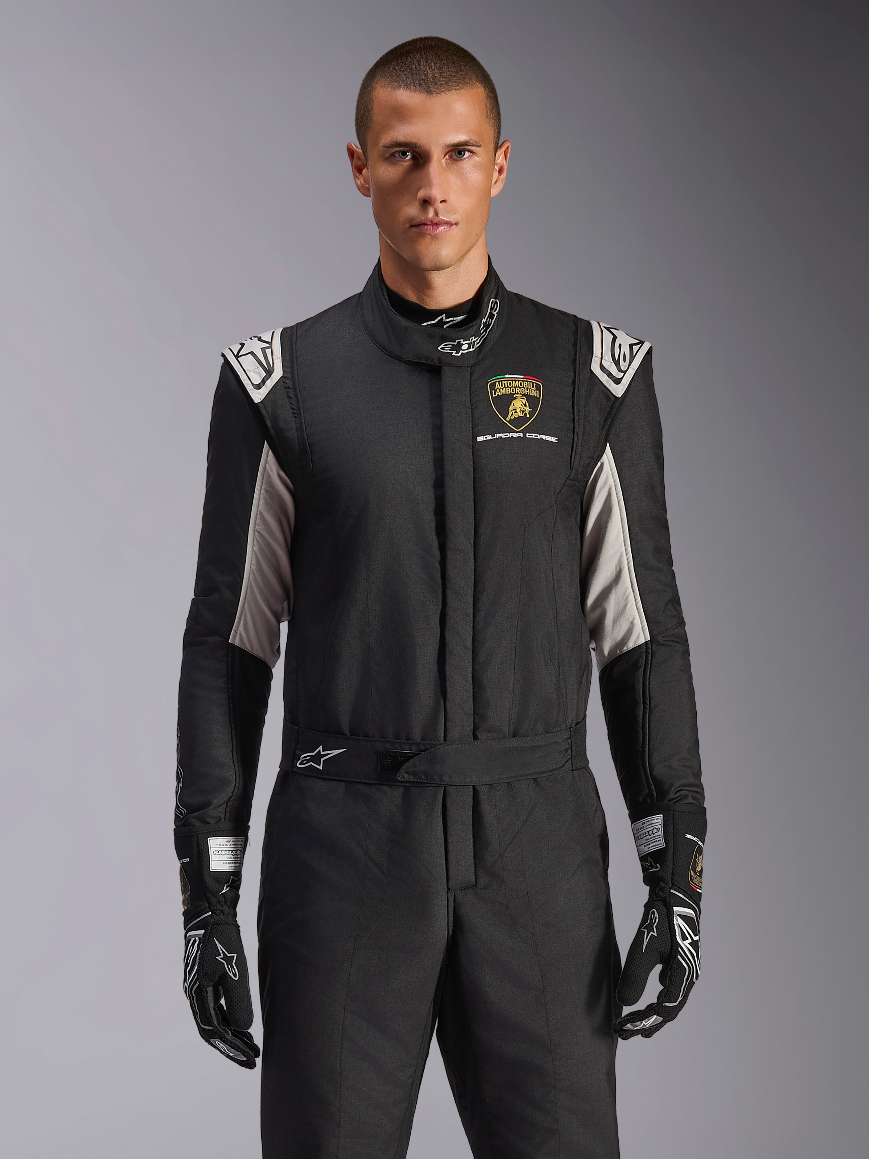Combinaison Alpinestars GP Tech V4 Édition Limitée Lamborghini, combinaison de course professionnelle noire avec accents argentés et dorés, arborant l'écusson Automobili Lamborghini Squadra Corse sur la poitrine, portée par un mannequin avec des gants de course noirs assortis.