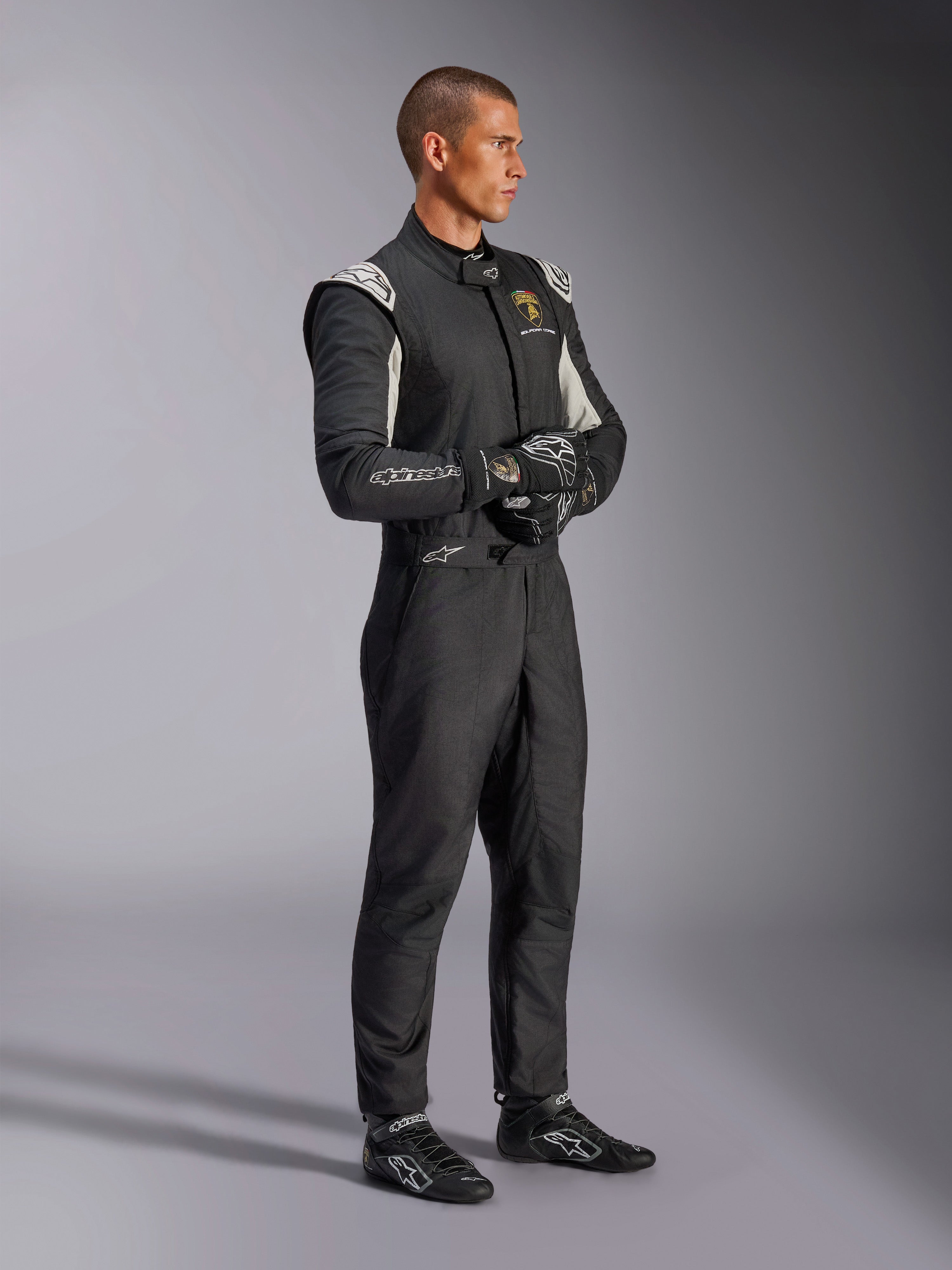 Pilote portant la combinaison Alpinestars Édition Limitée Lamborghini GP Tech V4, combinaison de course automobile certifiée FIA en noir, argent et or, avec gants et chaussures de course assortis, marquage Lamborghini Squadra Corse, équipement de sport automobile professionnel
