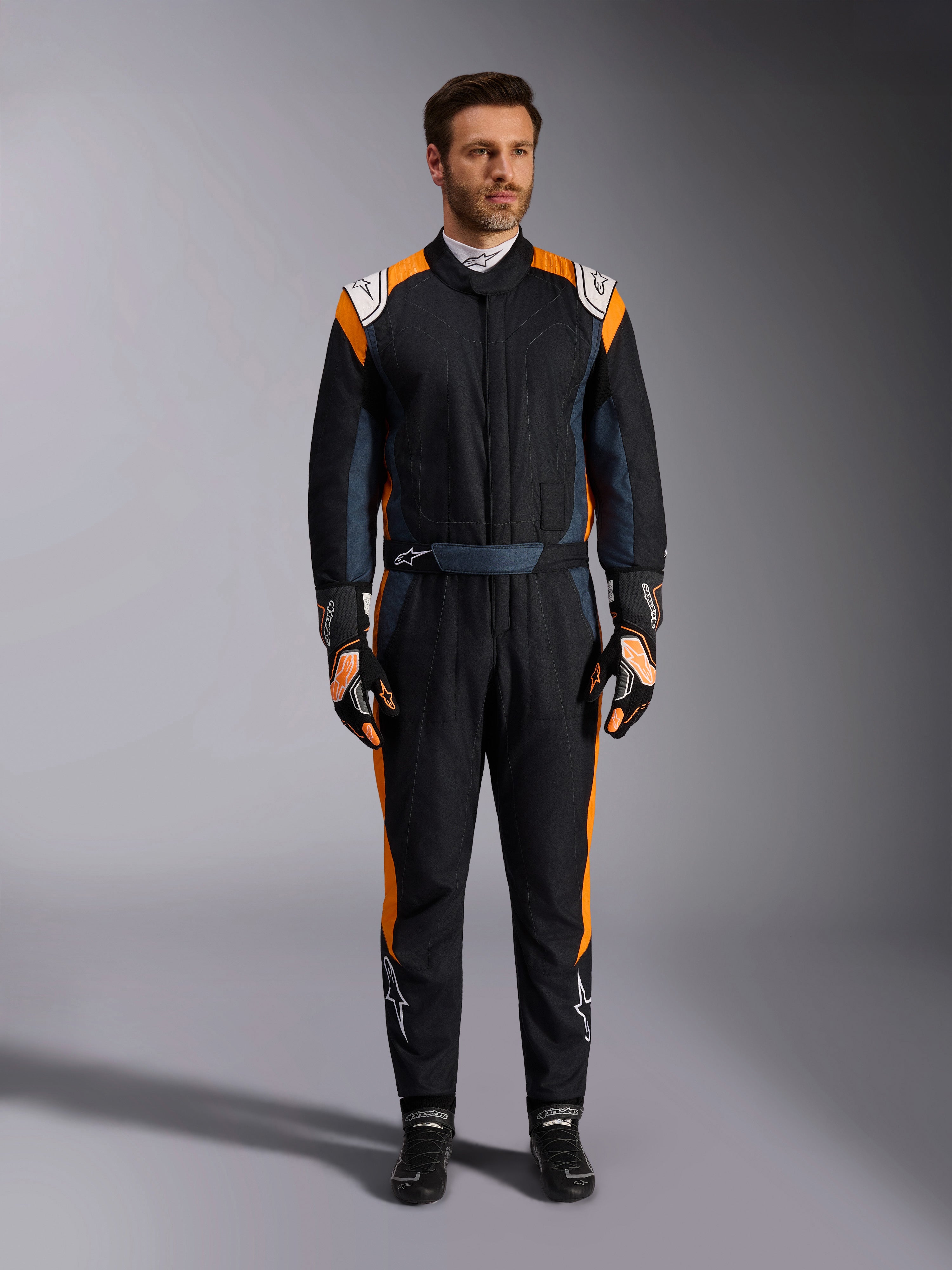 Combinaison Alpinestars GP Pro Comp V2 FIA, combinaison de course automobile noir asphalte et orange fluo, mannequin portant un équipement de protection complet avec gants et chaussures de course assortis, logos Alpinestars blancs et panneaux latéraux orange vif, vêtement de sport automobile professionnel