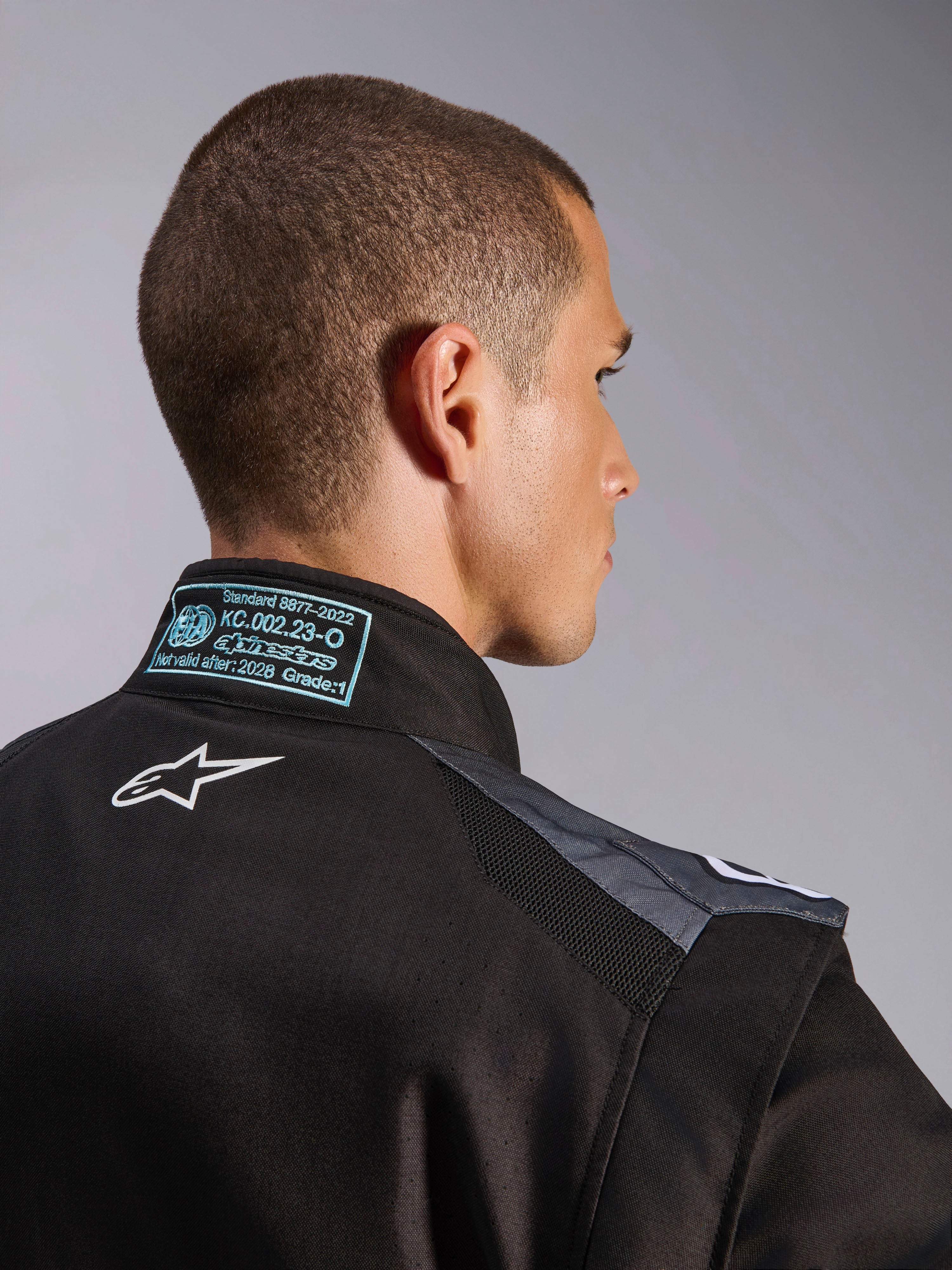 Alpinestars KMX-5 V3 Suit in Black and Dark Shadow, close-up view of the back and collar showing the white Astars logo, FIA 8877-2022 Grade 1 certification tag, and ventilated mesh shoulder panels.