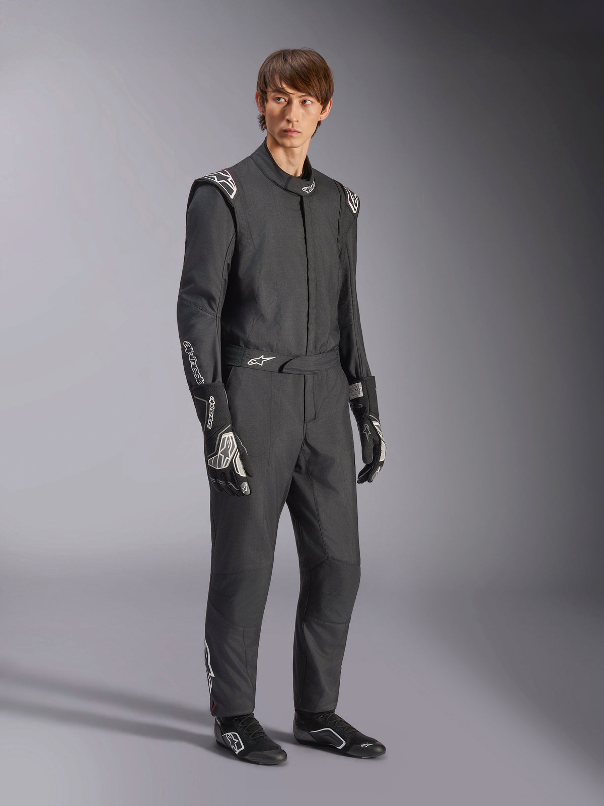 Alpinestars GP Tech V4 Suit, combinaison de course automobile une-pièce, noir et blanc, portée avec des gants de course et des chaussures assortis, arborant des logos blancs sur les épaules et les avant-bras, design professionnel épuré pour le sport automobile
