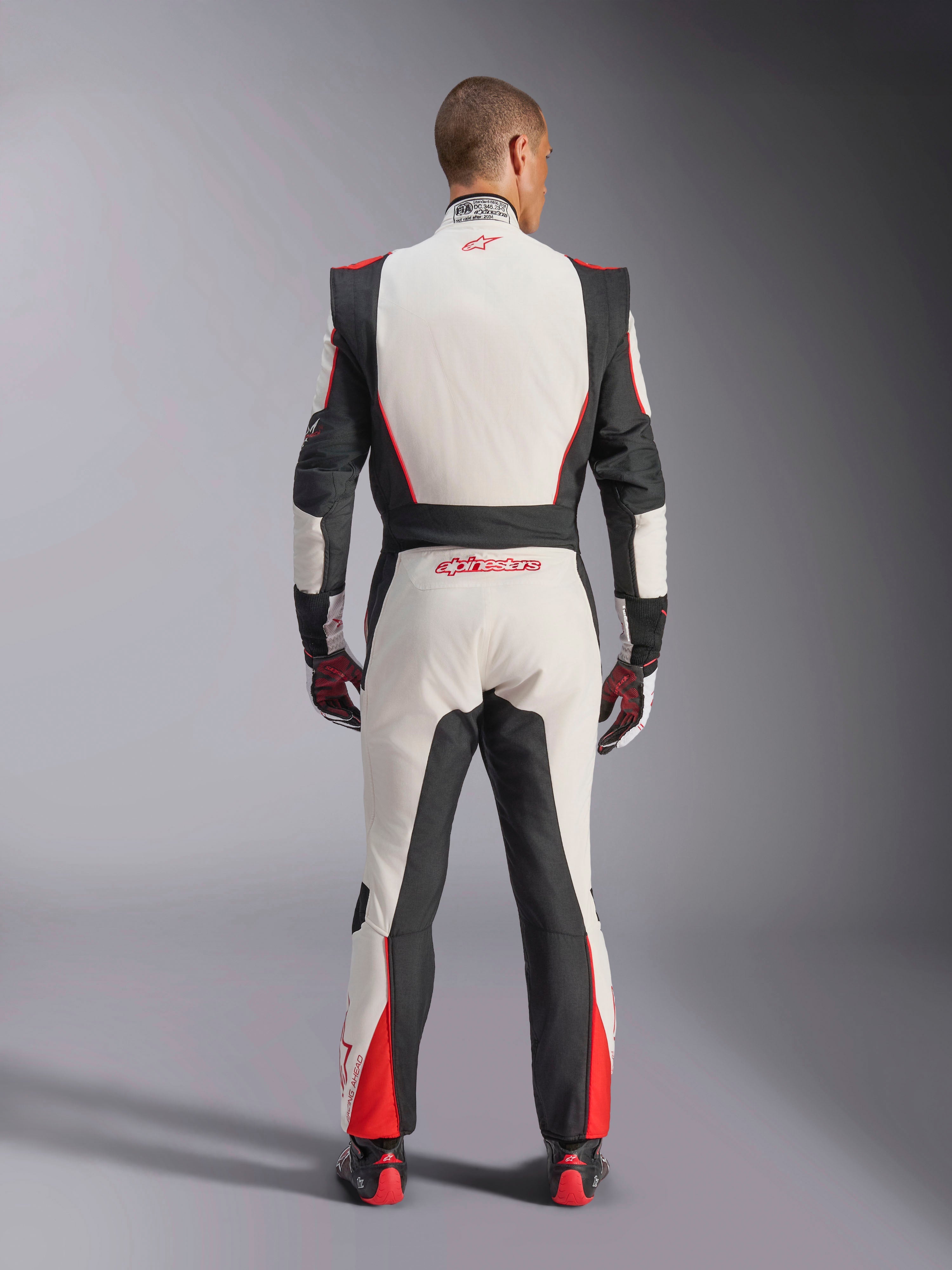 Alpinestars GP Tech V4 Suit, Auto Racing Suit, White Black and Red, back view of driver wearing the one-piece suit, featuring black stretch panels on the sides and shoulders, red accents on the lower legs, Alpinestars branding on the lower back and collar, professional FIA-certified motorsport gear
