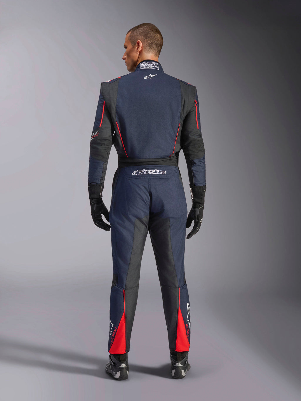 Vue arrière de la combinaison Alpinestars GP Tech V4 en bleu marine, noir et rouge, combinaison de course automobile une pièce avec panneaux extensibles noirs et accents rouges, logo Alpinestars et détails de certification FIA, portée avec gants et bottines de course professionnels.