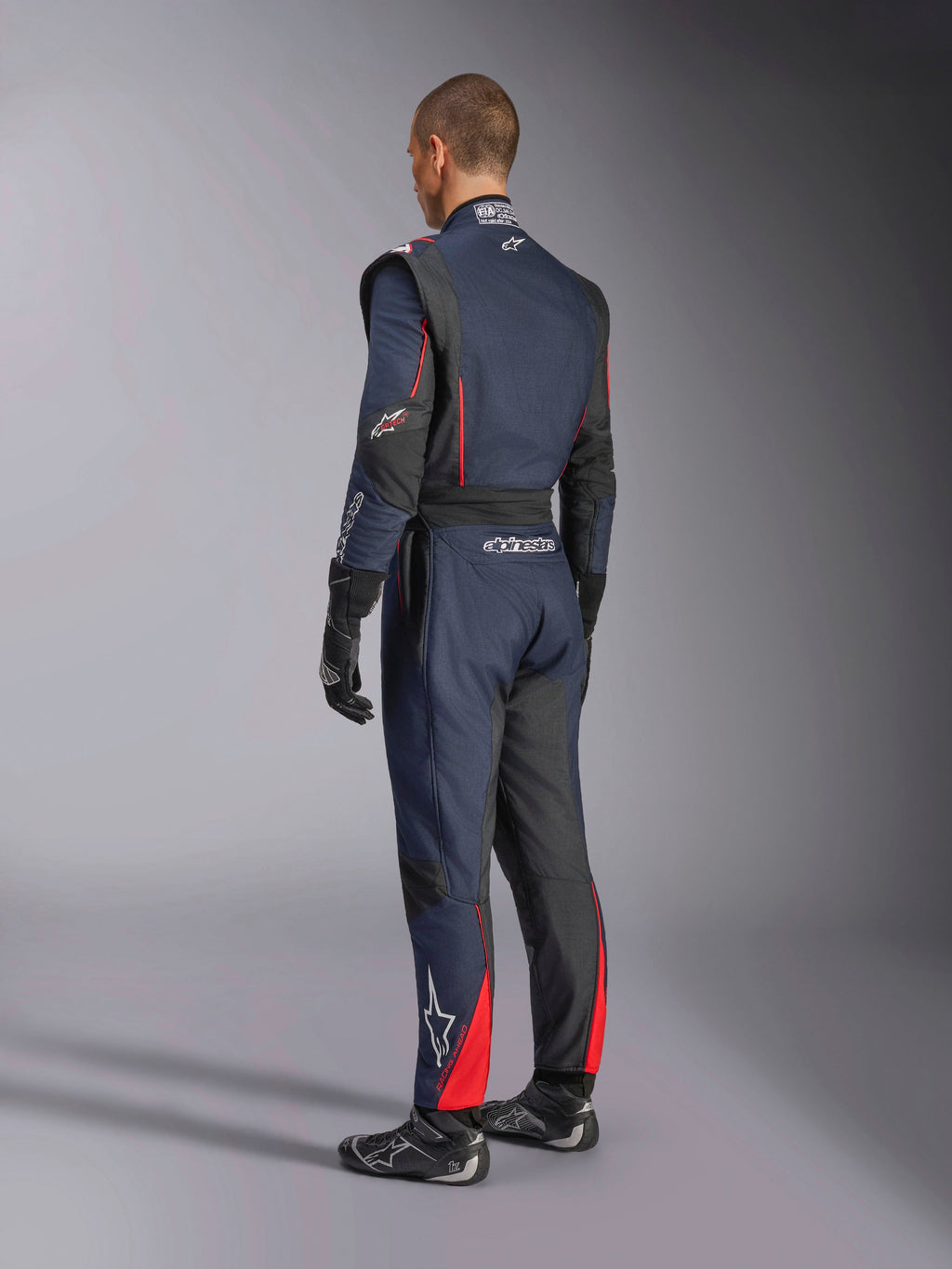 Combinaison Alpinestars GP Tech V4, Bleu Marine Noir Rouge, vue arrière latérale d'une combinaison de course automobile professionnelle une pièce, avec des accents rouges et des panneaux noirs, portée avec des gants et des chaussures de course noirs, équipement de sport automobile certifié FIA.