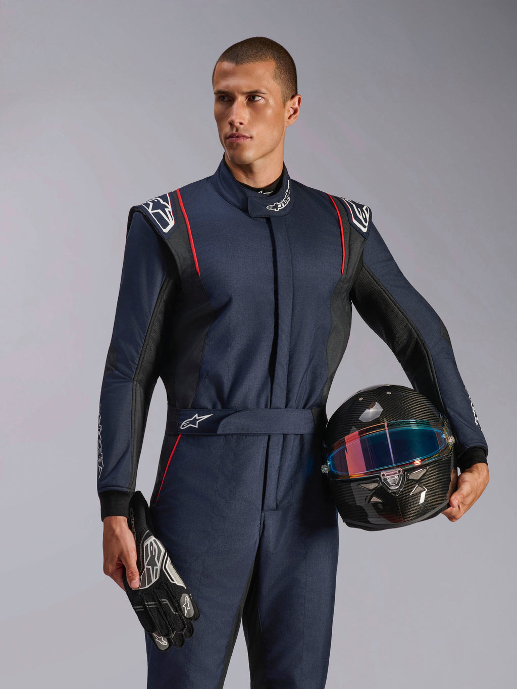 Mannequin homme portant la combinaison Alpinestars GP Tech V4, combinaison de course automobile bleu marine avec accents noirs et rouges, tenant un casque en fibre de carbone et des gants de course, équipement de protection professionnel pour le sport automobile