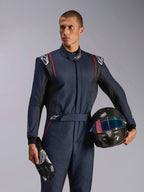 Mannequin homme portant la combinaison Alpinestars GP Tech V4, combinaison de course automobile bleu marine avec accents noirs et rouges, tenant un casque en fibre de carbone et des gants de course, équipement de protection professionnel pour le sport automobile