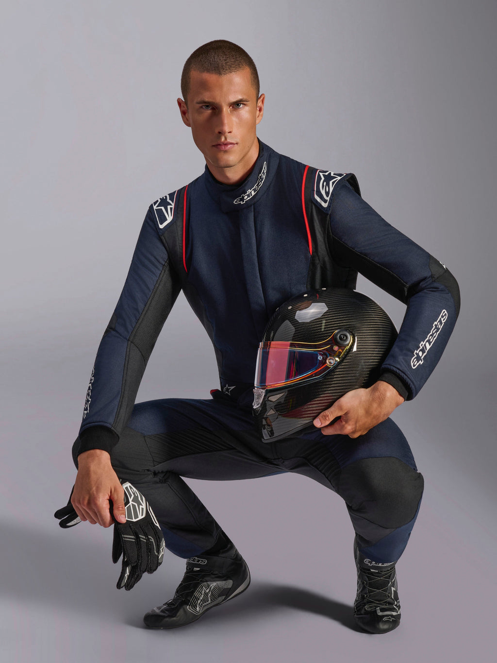 Pilote de course portant une combinaison Alpinestars GP Tech V4, bleu marine avec des accents noirs et rouges, tenant un casque en fibre de carbone avec une visière irisée, gants et bottes de course noirs, équipement de course automobile professionnel