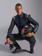 Pilote de course portant une combinaison Alpinestars GP Tech V4, bleu marine avec des accents noirs et rouges, tenant un casque en fibre de carbone avec une visière irisée, gants et bottes de course noirs, équipement de course automobile professionnel