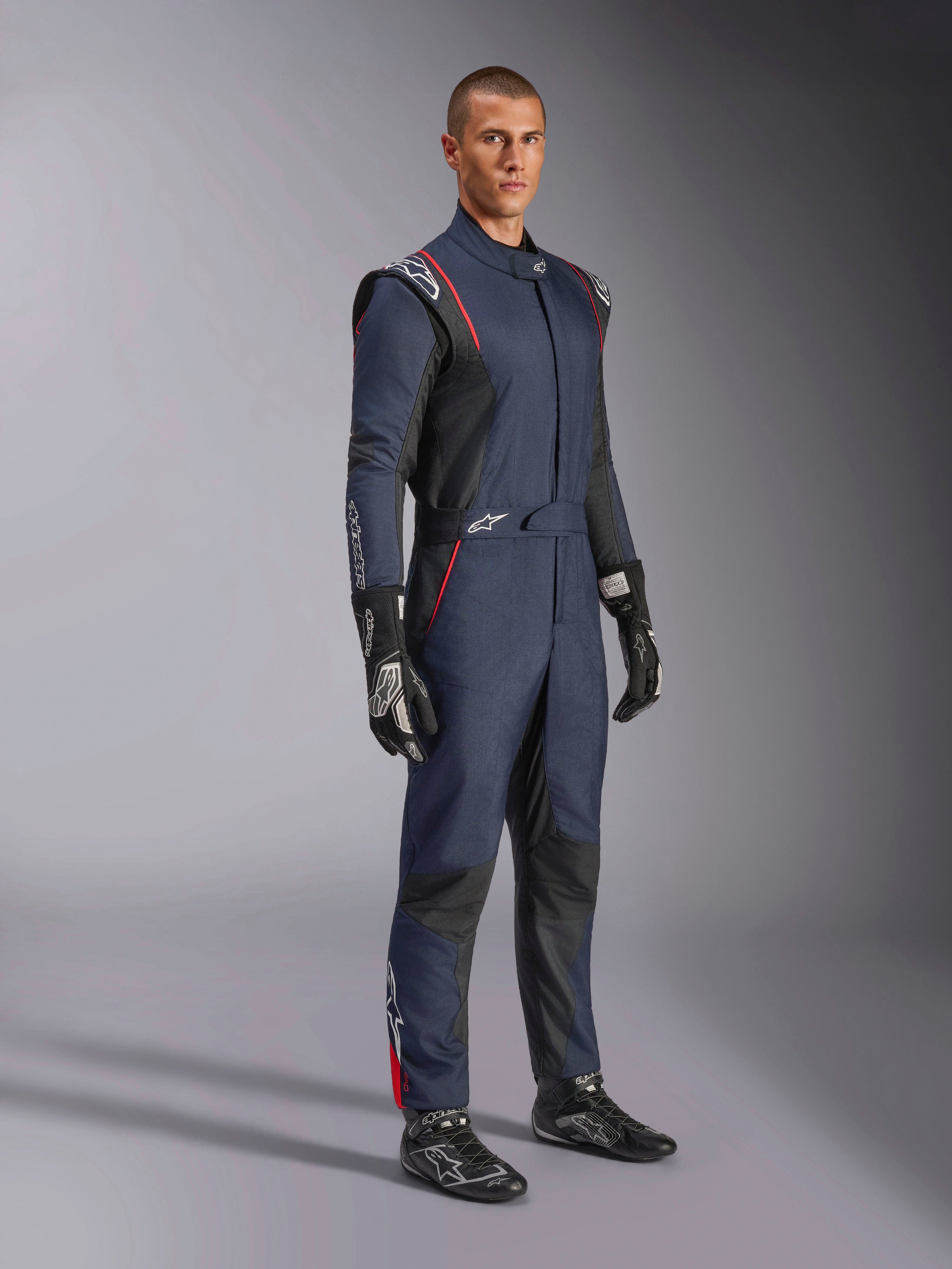 Combinaison Alpinestars GP Tech V4, combinaison de course automobile une pièce, bleu marine, noir et rouge, portée par un mannequin avec des gants et des bottines de course noirs, dotée de panneaux noirs et de liserés rouges, équipement de sport automobile professionnel.
