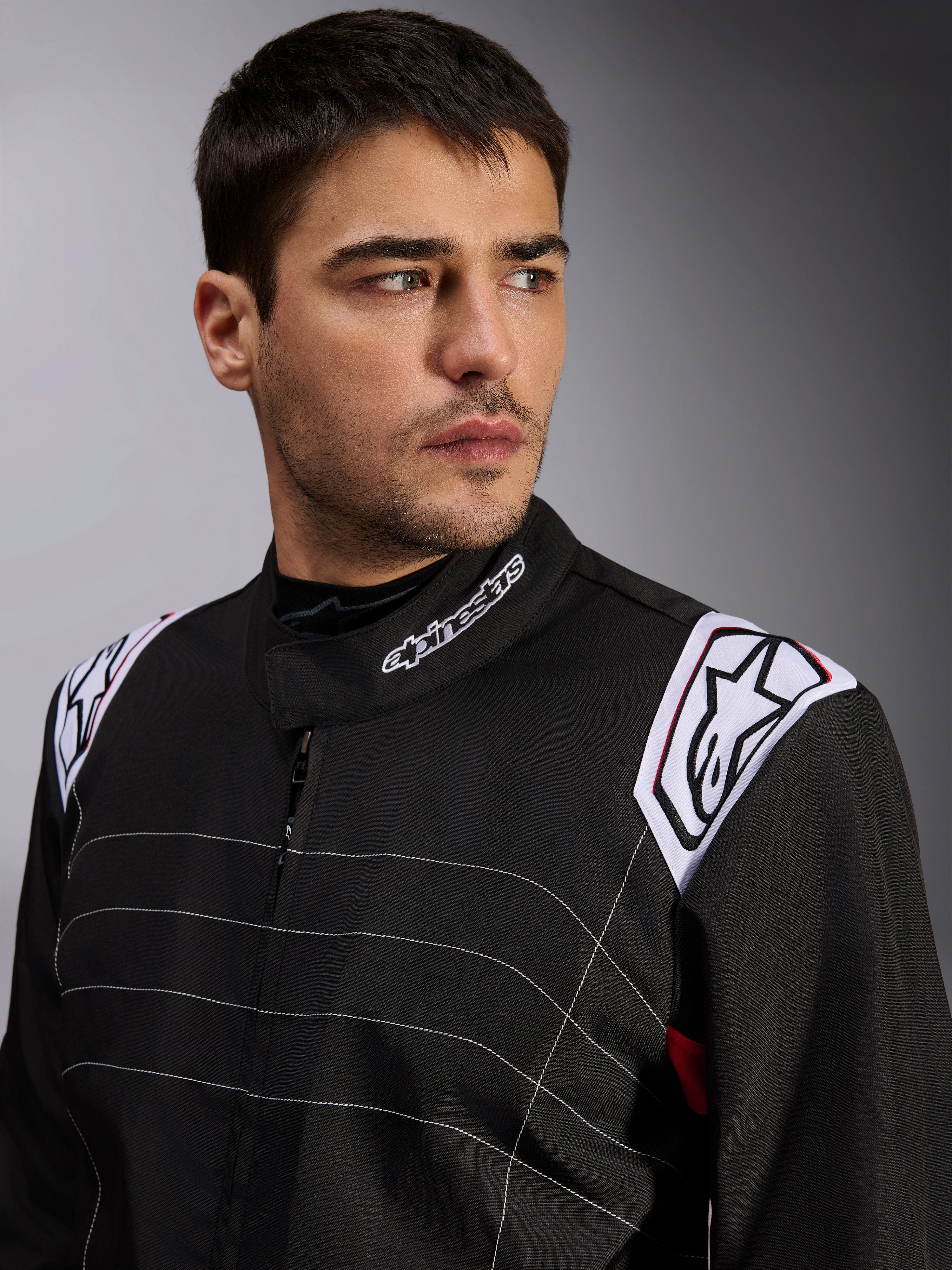 Alpinestars KMX-9 V3 Suit, Black Karting Suit worn by a male model, close-up of chest and shoulders with white branding on the collar and epaulettes, featuring white decorative stitching and a professional motorsport design