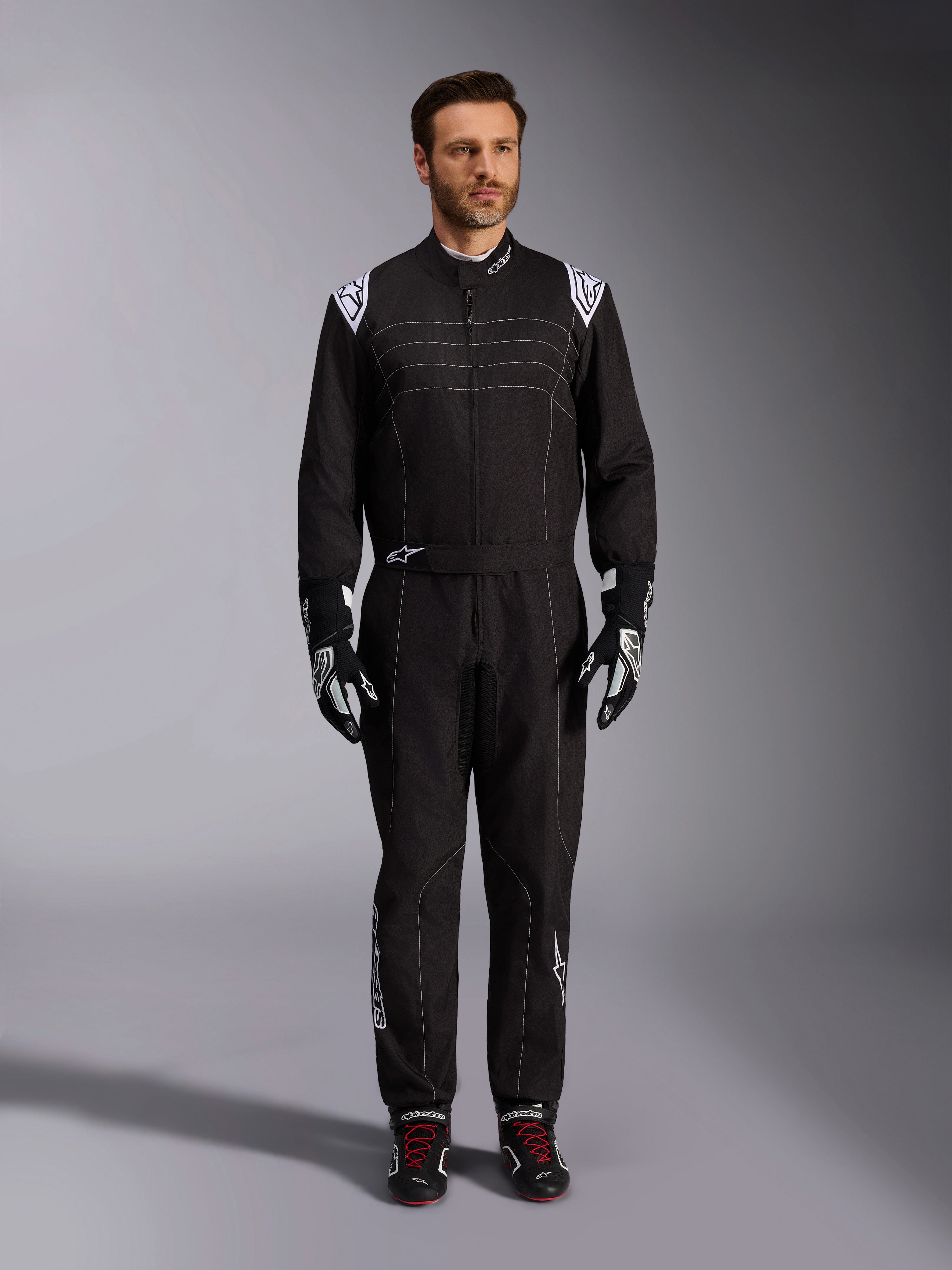 Combinaison Alpinestars KMX-9 V3, combinaison de karting noire et blanche, vue en pied sur un mannequin, avec des logos et des accents blancs sur les épaules, assortie de gants de course noirs et blancs et de chaussures noires avec des lacets rouges, coupe sportive-régulière pour équipement de karting professionnel.