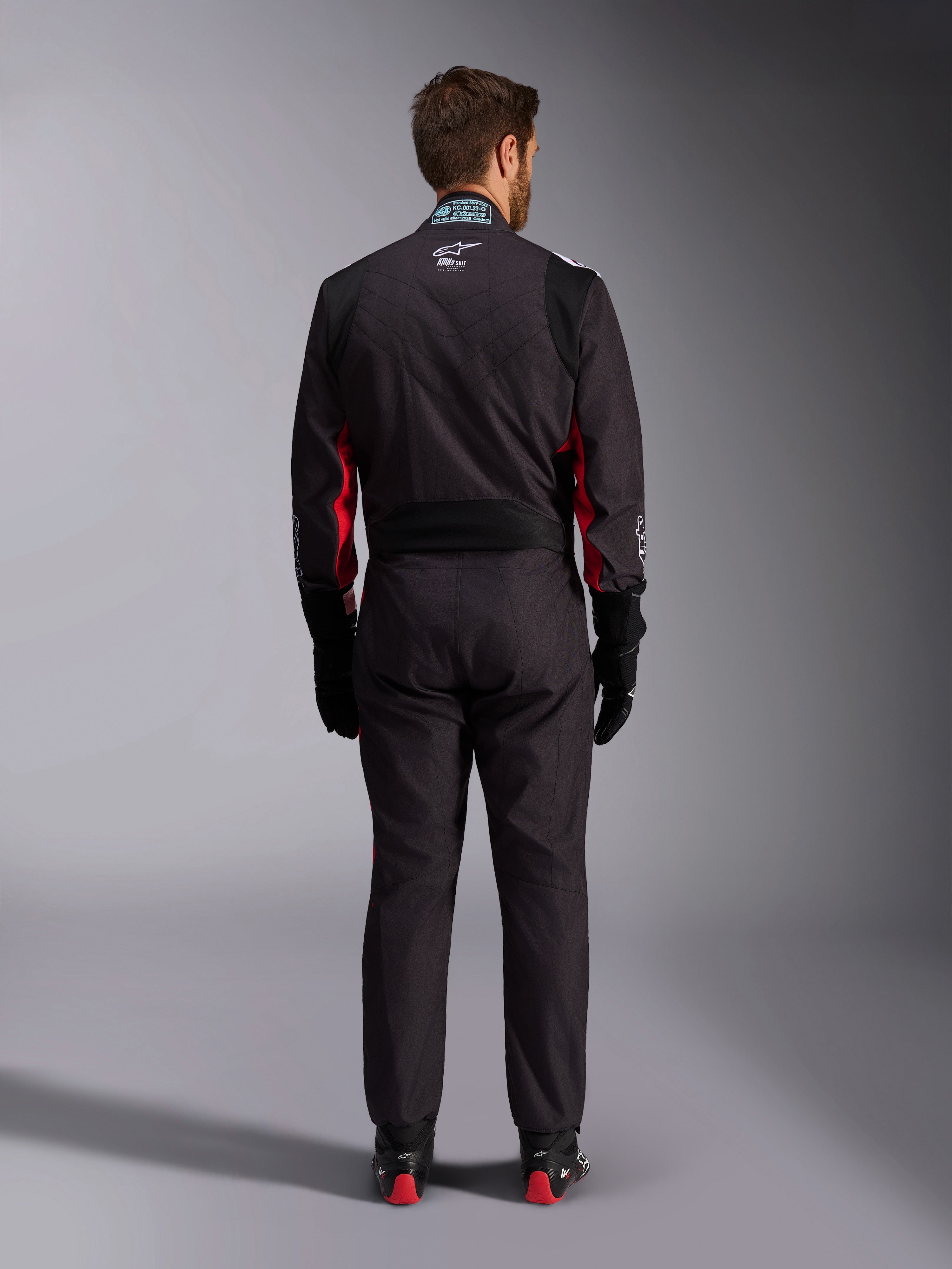 Combinaison Alpinestars KMX-9 V3 Graphic 3, combinaison de karting, noir et rouge, vue arrière présentant le panneau extensible lombaire et l'étiquette d'homologation FIA sur le col, accompagnée de gants de course noirs et de bottes aux accents rouges