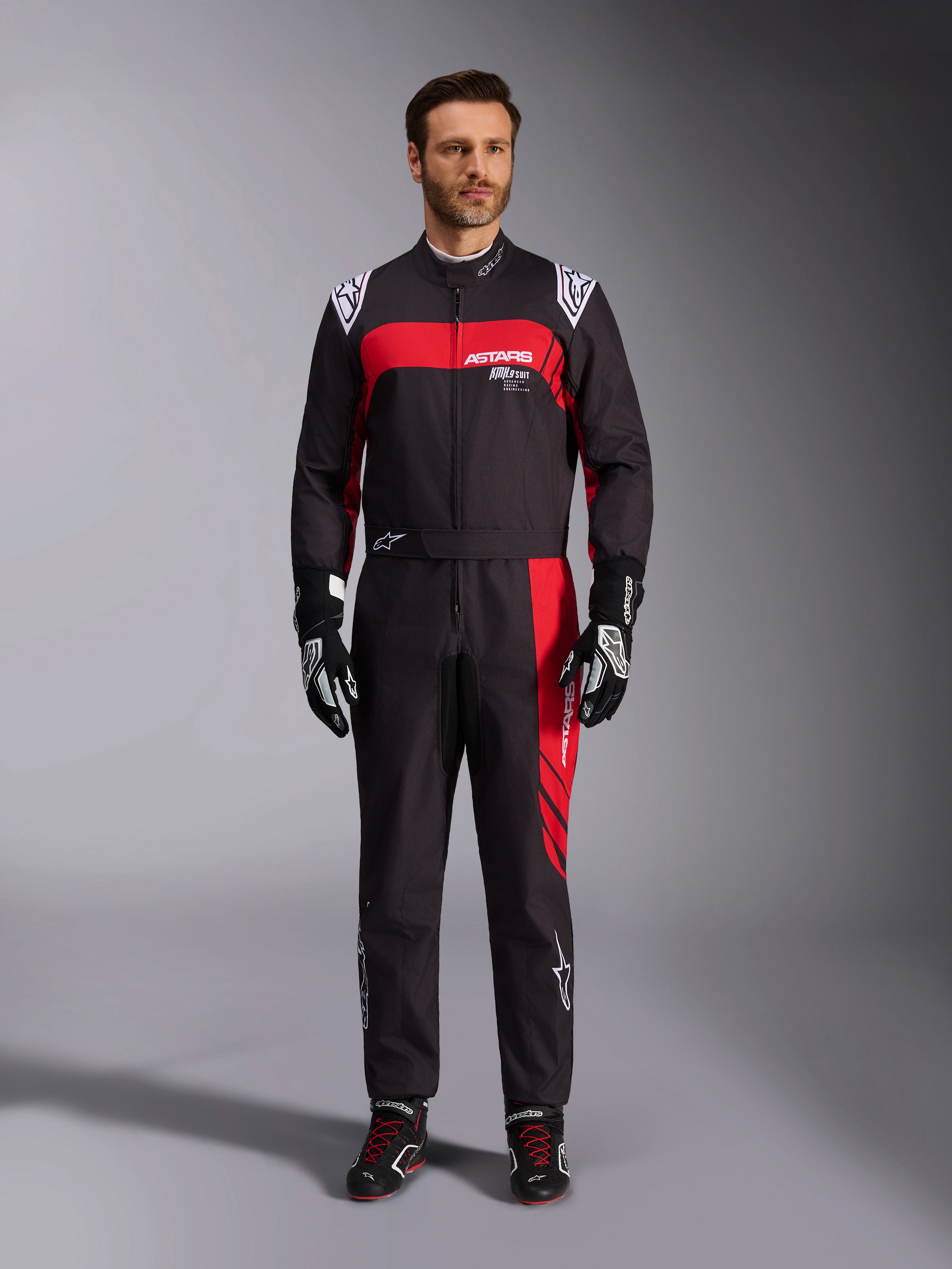 Combinaison Alpinestars KMX-9 V3 Graphic 3, combinaison de karting, noir et rouge, vue frontale en pied portée par un mannequin, avec des empiècements graphiques rouges et des logos blancs, associée à des gants et des chaussures de course noirs, équipement de sport automobile professionnel.