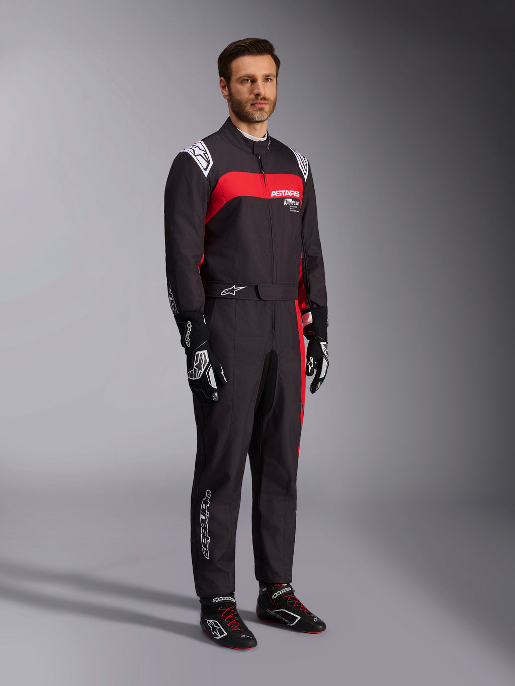 Mannequin homme portant la combinaison Alpinestars KMX-9 V3 Graphic 3, combinaison de karting, noir et rouge, avec épaulettes blanches et logo rouge sur la poitrine, gants de course noirs et chaussures à lacets rouges, équipement professionnel de sport automobile.