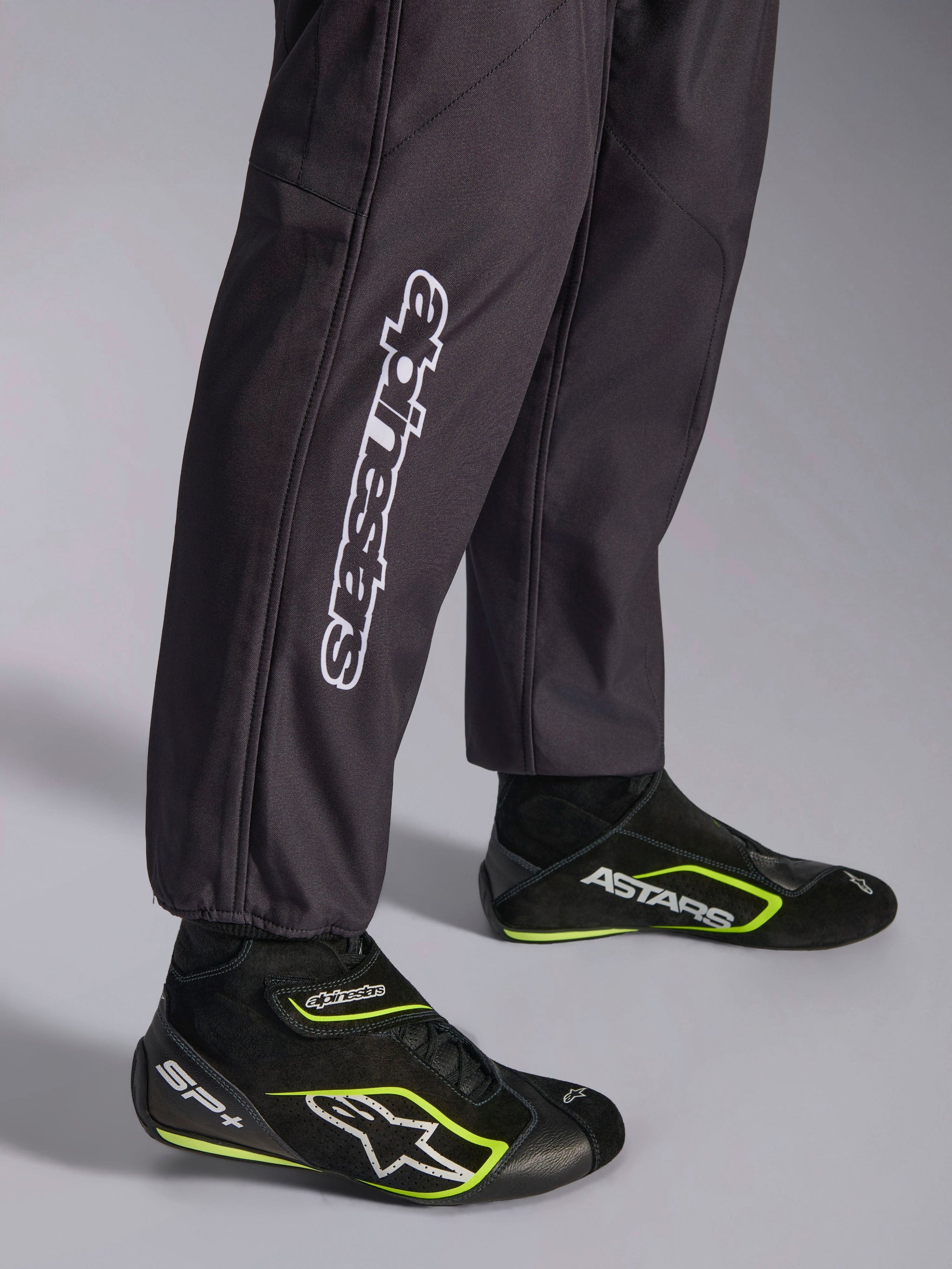 Alpinestars KMX-9 V3 Graphic 3 Suit, Karting Suit, Black, close-up of the lower legs and ankle cuffs, featuring white vertical Alpinestars branding, paired with black and yellow fluo karting shoes, detail of professional karting apparel.