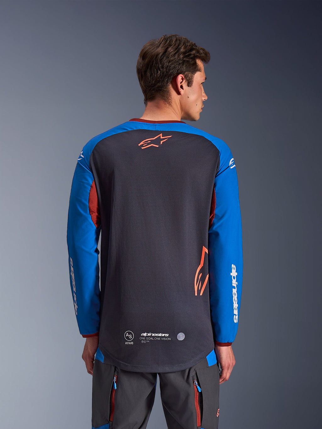 Maillot Alpinestars Maxdura Dual en gris foncé et bleu royal, vue de dos d'un modèle masculin, torse en maille avec logo Astars orange, manches longues avec marquage blanc, col et poignets bordeaux, porté avec un pantalon technique gris, équipement de moto dual-sport et d'aventure